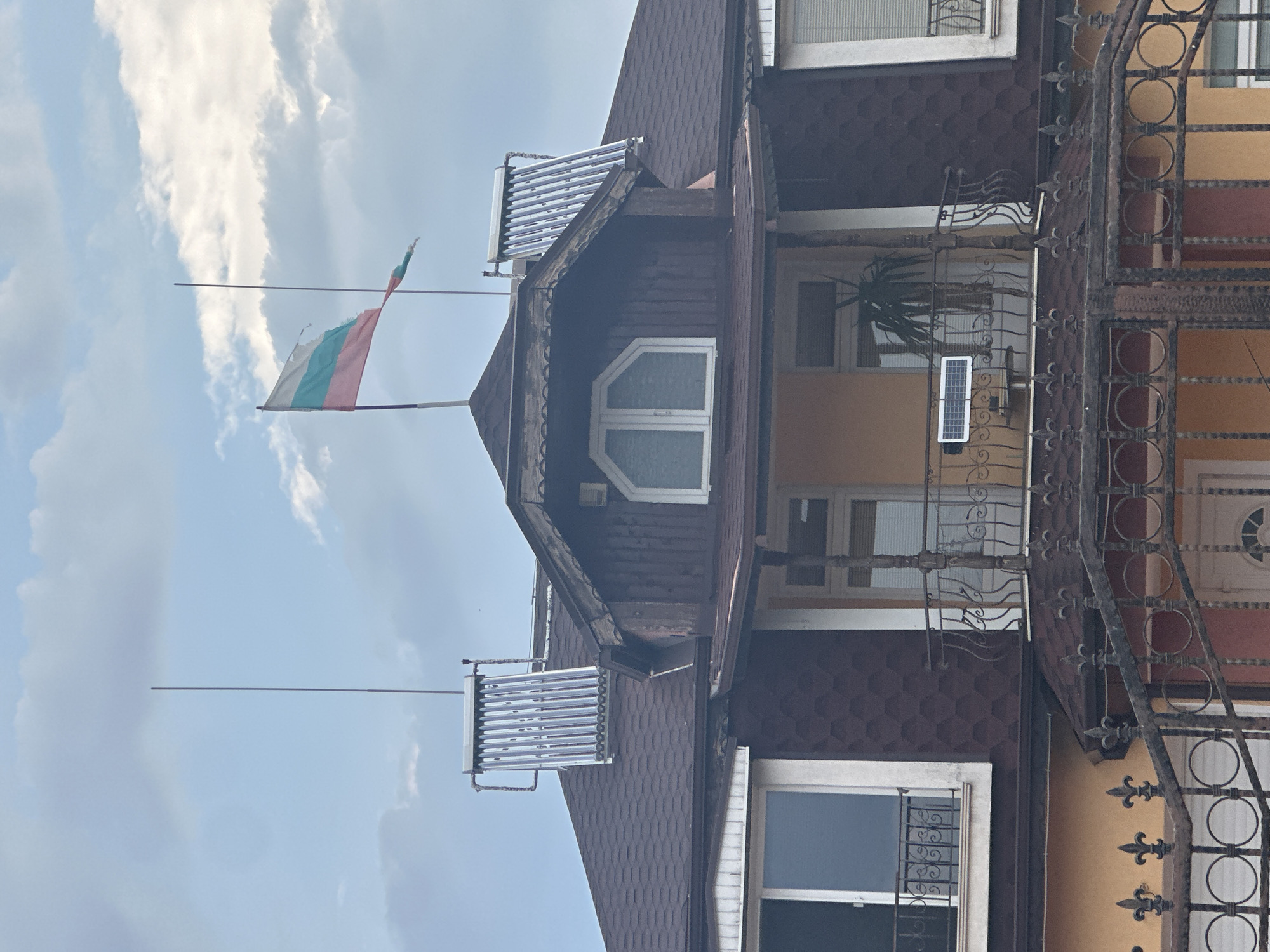 The image shows the upper facade of a building with a brown shingled roof. There are two solar water heaters mounted on the roof. A Bulgarian flag flies from a pole above the roofline. Below the flag is a small balcony with a wrought-iron railing. A small solar panel is mounted on the balcony railing, and a potted plant is visible on the balcony. Two windows with wrought-iron balconies are visible on the lower part of the facade. A dark metal fence with ornate finials runs across the bottom of the image, in front of an orange-colored wall with a white door. The scene is set against a cloudy sky. The location is identified as Kashin, Bulgaria.