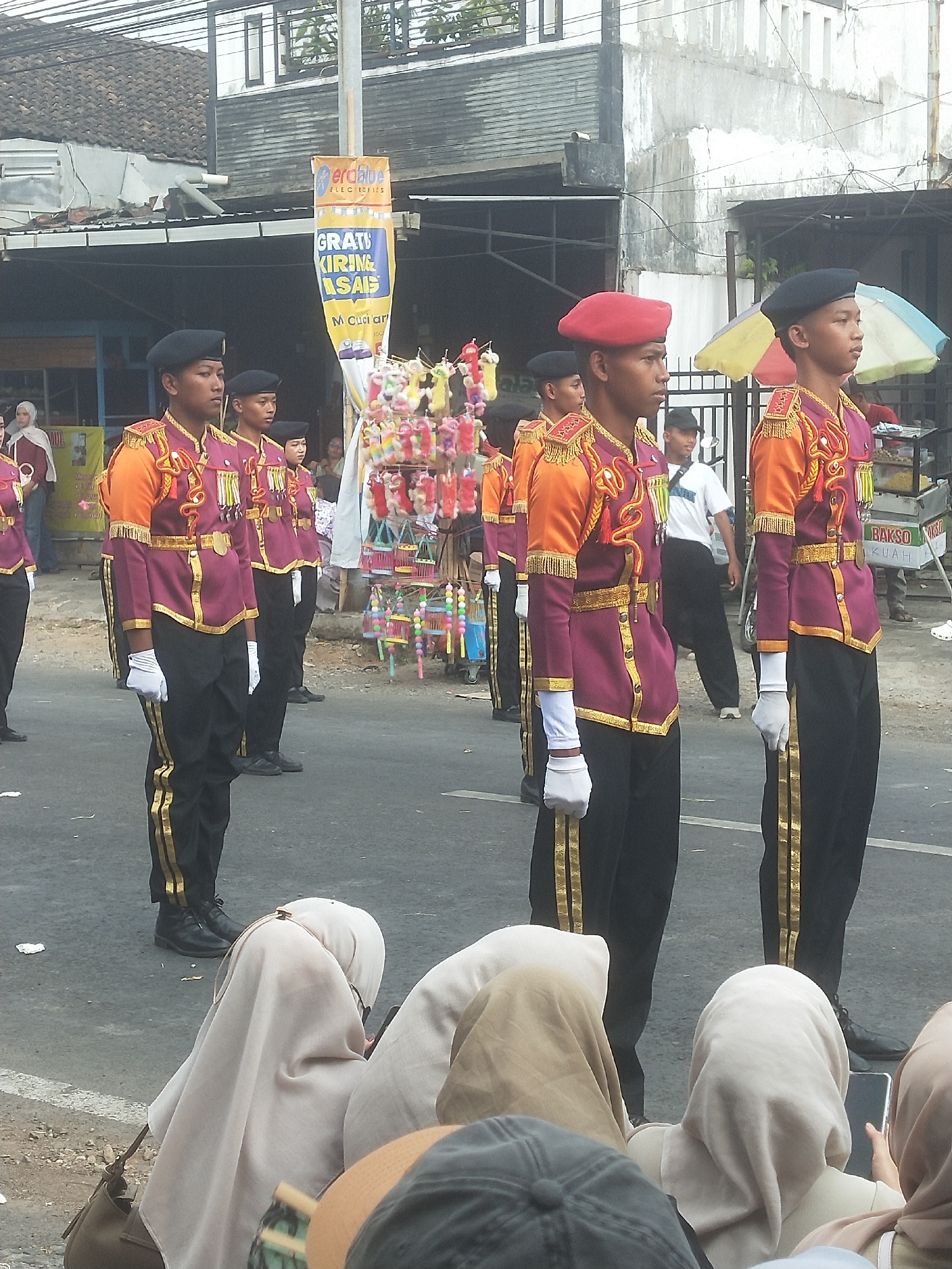 The image depicts a parade in Boja, Indonesia, with a group of young men in marching band uniforms. The men are wearing maroon jackets with gold trim and epaulets, orange sleeves, black pants with gold stripes, and black hats. Some are wearing white gloves. They are standing in a line on a street, with spectators in the foreground wearing hijabs. In the background, there are buildings, a banner advertising "ERAbhup Electronics" and "GRATIS KIRIM ASAS", and stalls selling snacks and toys. The weather appears to be overcast.