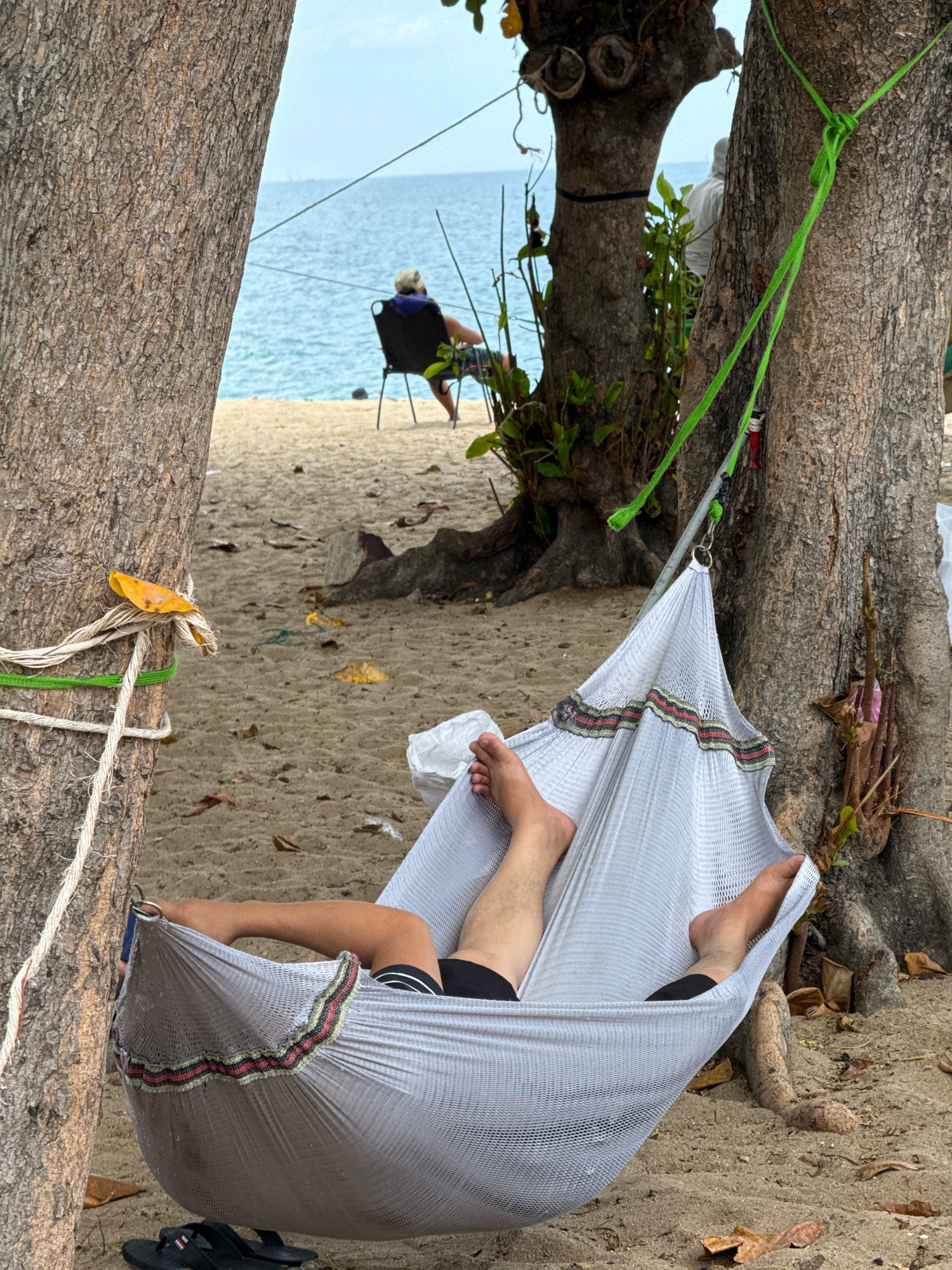 The image depicts a serene beach scene in Nha Trang, Vietnam, featuring two individuals relaxing by the ocean. In the foreground, a person is comfortably suspended in a white mesh hammock, their legs visible and bare, suggesting a warm climate. The hammock is strung between two large trees on a sandy beach, with fallen leaves scattered on the ground. A pair of black flip-flops rests on the sand near the hammock. In the middle ground, another person is seated in a dark chair, facing the ocean, their back to the camera. They are wearing a hat and appears to be enjoying the view. The ocean stretches out in the background, with a clear blue sky overhead. The overall atmosphere is one of relaxation and leisure, characteristic of a tropical beach destination.