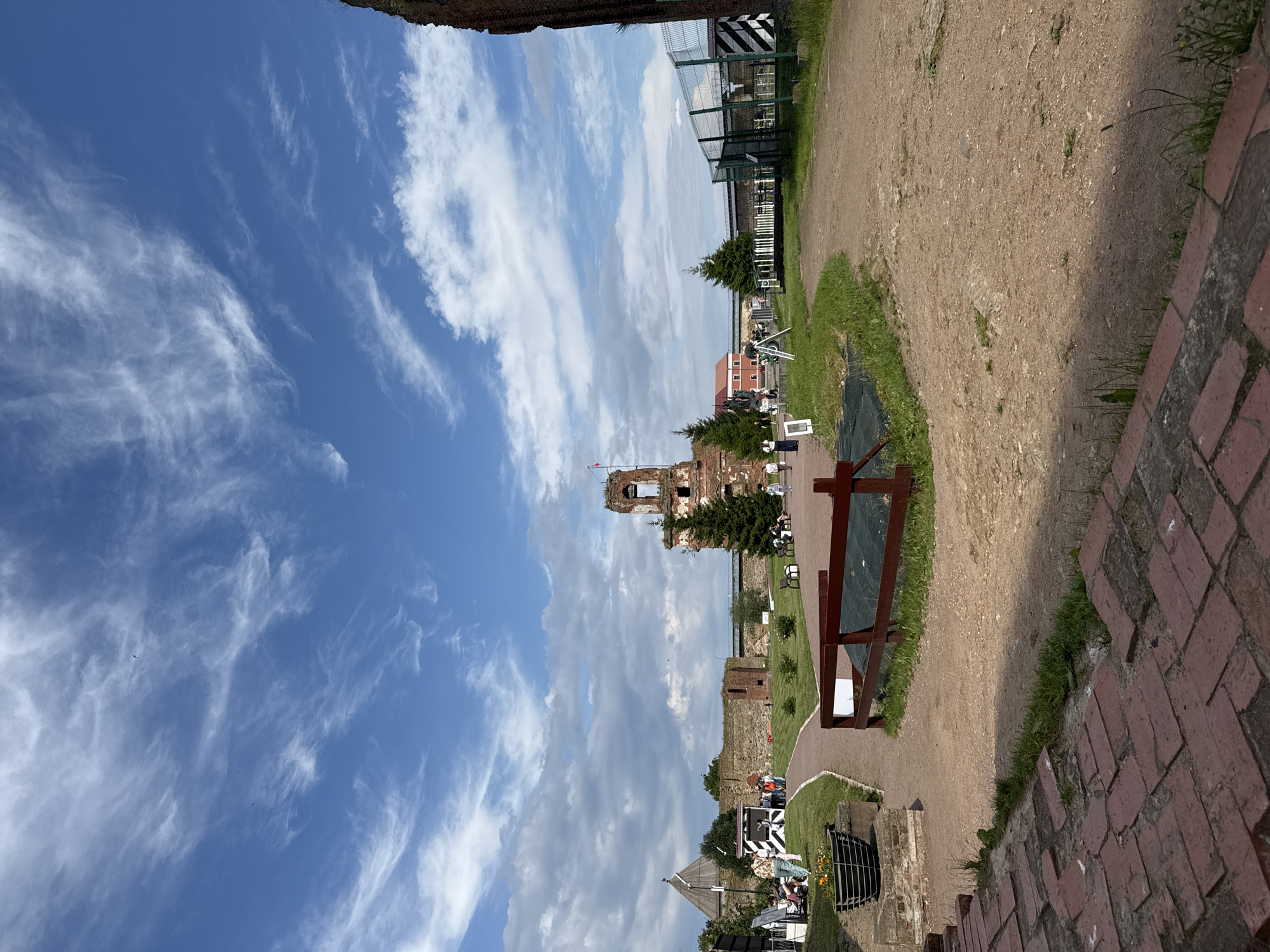The image captures a view of the Oreshek Fortress, located on an island in the Neva River, Shlisselburg, Russia. The scene is dominated by a bright blue sky with scattered white, wispy clouds. In the foreground, a brick pathway leads to a dirt path that curves through a grassy area. Several people are visible, some sitting on benches, others walking. In the midground, the fortress walls, constructed of stone and brick, enclose the grounds. A prominent feature is a crumbling brick tower, its upper section open to the sky, with some greenery growing around its base. Several evergreen trees are also scattered throughout the fortress grounds. A metal fence with black and white striped barriers runs along the right side of the image, defining the edge of the area. The overall impression is of a historical site on a sunny day with visitors exploring the grounds.