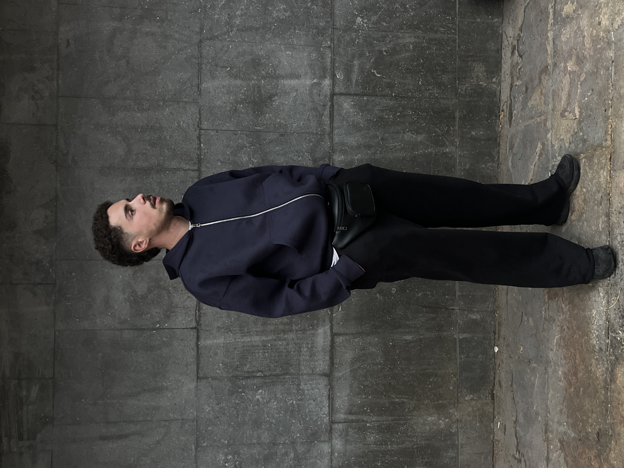 A young man is depicted lying flat on a textured, tiled ground surface, looking upwards. The photograph is oriented vertically, with the man positioned horizontally across the frame, occupying the lower and central part of the image.
The subject has dark, curly hair and a short beard or stubble. He is dressed in a dark navy blue zippered jacket or sweatshirt, black trousers, and dark, flat-soled shoes. A black fanny pack or small utility bag is worn around his waist, positioned over his jacket. A silver earring is visible in his left ear.
The setting is an outdoor urban environment, likely in Barcelona, Spain, given the context. The ground is paved with large rectangular tiles that show signs of wear and varying shades of grey and brown, with some lighter patches indicating dust or dirt. A distinct vertical line (when the image is viewed in its current rotated state) separates a darker, more uniform grey tiled area from a lighter, more weathered brownish-grey section. The lighting suggests it is daytime, but no harsh shadows are present, indicating either an overcast sky or a shaded location. The man's expression is neutral as he gazes upwards, conveying a sense of contemplation or relaxation. No specific activity is taking place beyond this pose. There is no visible text within the image.