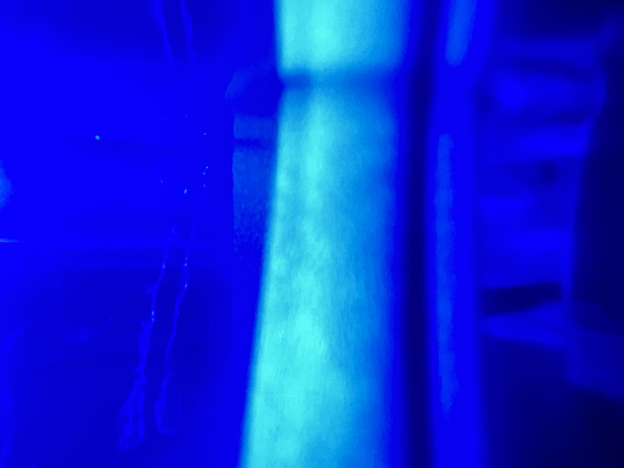 The media file is an abstract, blurry photograph dominated by shades of vibrant blue and a lighter cyan. The image appears to be taken in a dimly lit environment, possibly underwater or in a space with colored lighting, given the deep blue hues. There are no discernible people or specific objects, but abstract shapes and light patterns suggest a possibly dynamic or fluid scene. A lighter, almost vertical band of cyan light cuts through the center of the frame, creating a strong contrast with the surrounding blue. This light source seems to illuminate something, causing a hazy, textured effect within its glow. On the left side of the image, there are faint, wispy white or very light blue streaks, which could be interpreted as organic forms, currents, or debris. The overall impression is one of depth and perhaps mystery, with the bright cyan light acting as a focal point against the deep blue backdrop. No text is visible. The location context of Letterkenny, Ireland, doesn't provide specific visual cues within the abstract nature of the photograph itself.