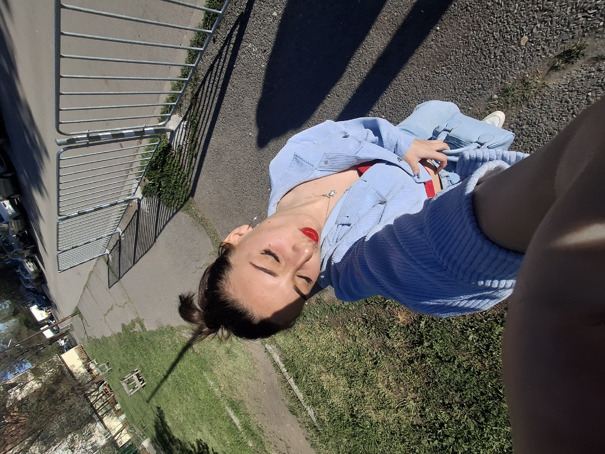 A woman takes a selfie outdoors. She wears a light blue corduroy jacket over a red garment and a necklace with a round pendant. She holds a light blue purse in her hands. She is wearing jeans and white sneakers. The background includes a fence, a paved area, grass, and trees, suggesting an urban park. Given the reference to Sofia, Bulgaria, the location could be a park or recreational area within the city.