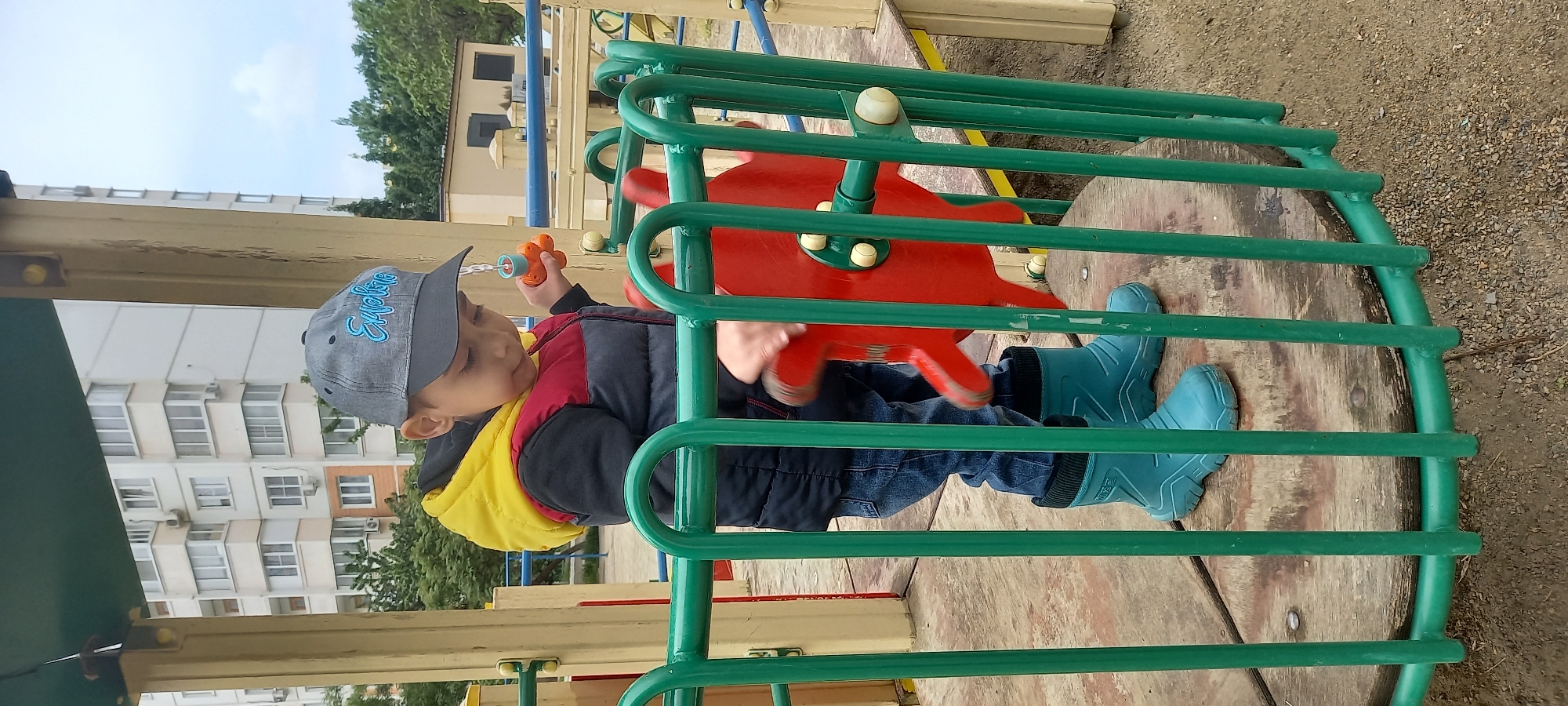 A child is playing on playground equipment in Krasnodar, Russia. The child, wearing a gray baseball cap, dark blue jacket with yellow lining, blue jeans, and teal rubber boots, is seated inside a green metal structure, holding a small orange and yellow toy in their left hand and turning a red wheel with their right hand. The playground equipment sits on a circular wooden platform. In the background are apartment buildings and trees. The ground surrounding the equipment appears to be gravel or dirt. The sky is visible and appears overcast.