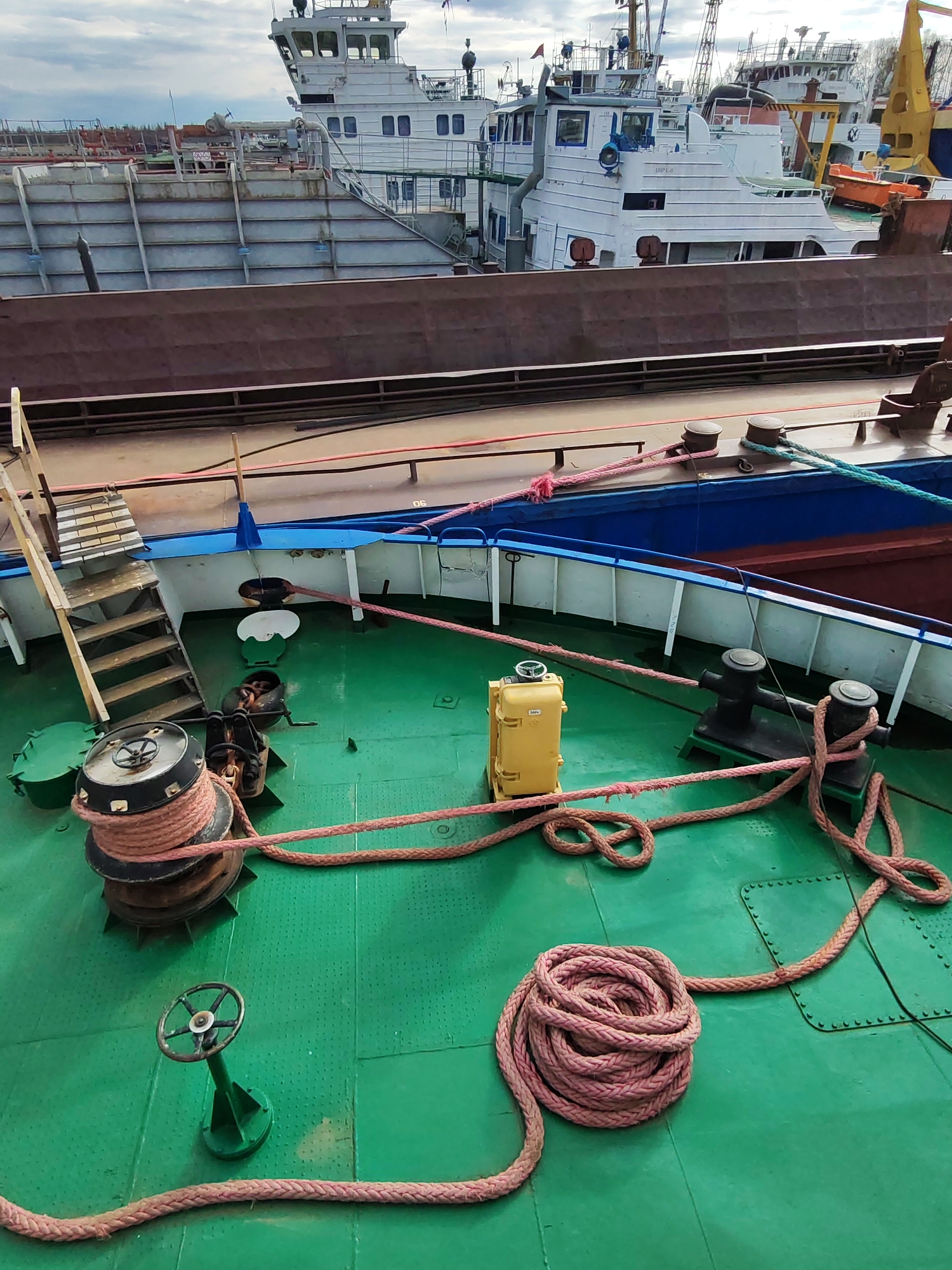 This image shows the deck of a boat in the city of Rybinsk, Russia, likely docked at a port. The deck is green and features various pieces of nautical equipment, including a large winch with a coil of rope, a black bollard with rope wound around it, and a smaller green wheel-operated device. There is a yellow box-like object nearby. A length of thick, pinkish-red rope runs across the deck in various loops and coils. In the background, other ships are visible, including a large, white vessel with a multi-tiered deckhouse and a brown-sided ship docked alongside. A set of wooden stairs leads up to another level of the ship. The sky is overcast.