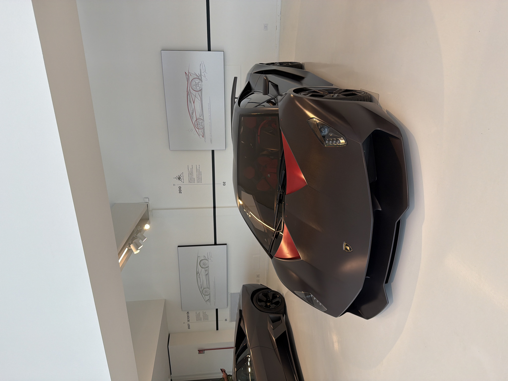 In a museum setting in Maggi, Italy, a matte gray Lamborghini Sesto Elemento is displayed on a white floor. The car features a prominent red accent on its hood and red interior seats. To the left of the Sesto Elemento, a dark gray Lamborghini Reventón is partially visible. The walls are white, with black accents and framed automotive design sketches. One sketch, labeled "2007 REVENTÓN," depicts the Reventón, and another, labeled "2010 SÉSTO ELEMENTO," shows a design for the Sesto Elemento. The museum is lit by overhead track lighting. A person, mostly obscured, is visible on the far left.