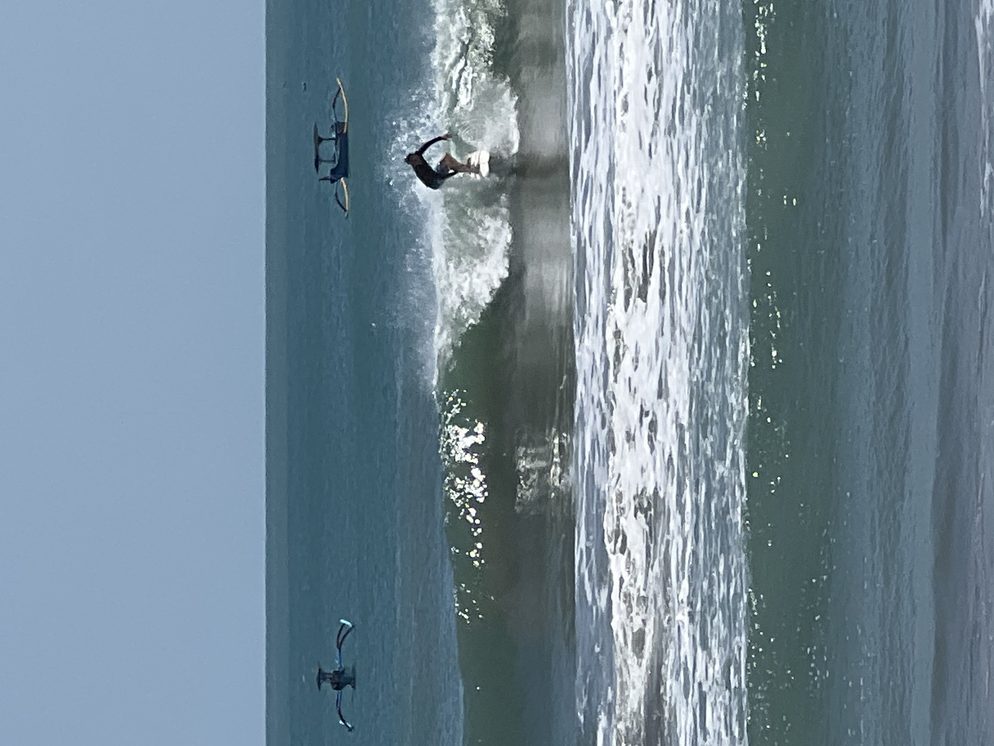 A surfer rides a wave on the ocean off the coast of Kuta, Indonesia. The surfer is a male, wearing dark shorts and a dark shirt. He is standing on a white surfboard, in the middle of a breaking wave. The ocean is a deep blue-green color, with white foam from the waves. In the background, there are two traditional Indonesian outrigger boats. One boat is on the left side of the image, and the other is on the right. The sky is clear and blue. The image appears to have been taken from the shore.
