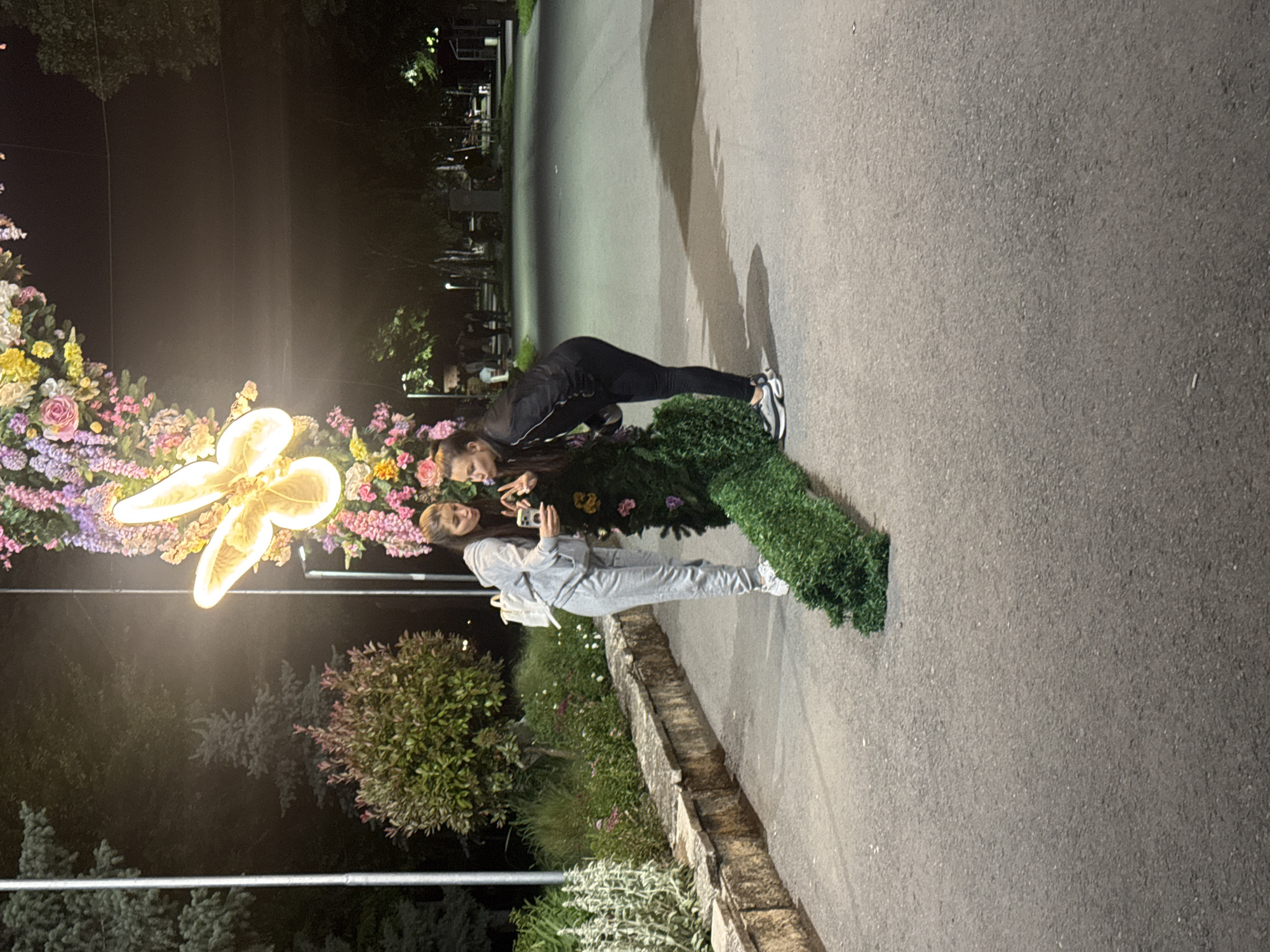 Two females pose for a photo in Shumen, Bulgaria. One in black workout attire bends over facing the other, who wears a gray sweatsuit and holds a phone to take a picture. Behind them is a floral display with a lit butterfly emblem. They stand on a sidewalk next to a raised stone planter with greenery, beside a dark paved road. The location is lit by street lights. Trees are visible in the background.