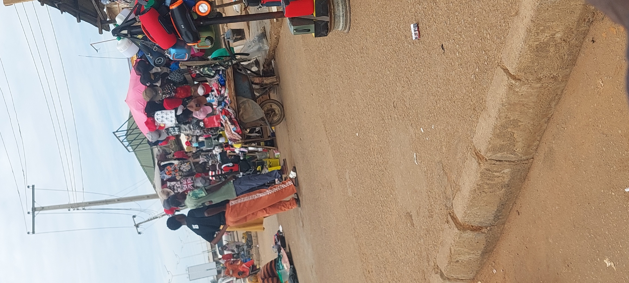 The image depicts an outdoor market scene in the city of Unknown, Unknown, under a cloudy daytime sky. The ground is a reddish-brown, textured asphalt or packed earth road, with a concrete curb visible in the foreground.
In the mid-ground, two dark-skinned men stand facing right, observing the market stalls. The man on the left wears a black short-sleeved shirt with a white number "15" on the back, orange track pants with a white checkered pattern down the sides, and white slide sandals. He stands with his hands on his hips. To his right, the second man wears a green short-sleeved t-shirt, dark blue pants, and brown sandals. A yellow garment or object is partially visible behind the first man. Further back on the left, indistinct figures and vehicles are present, suggesting a street or intersection. A person in a striped top is seated on the ground in the far left, beside items spread on a green tarp.
The right side of the image is dominated by crowded market stalls displaying merchandise. A large, open pink umbrella provides shade over one section. Numerous hats of various colors and designs, including baseball caps, bucket hats, some with cross motifs, and one with a Union Jack pattern, are densely hung and stacked. Various bags, including backpacks and shoulder bags, are also displayed. The stalls are covered by a green tarp roof supported by a wooden structure. A metal wheelbarrow filled with bundled textiles or clothing is parked near the front of the stalls. To the far right, a vertical support pole for a stall is visible, from which several items hang, including a red speaker and an orange speaker-like device with a light.
In the upper left, a light-colored utility pole with multiple horizontal power lines spans across the sky. Another utility pole is visible further in the background. Scattered small white debris and a small rectangular object with red and blue markings (possibly a cigarette pack) are visible on the road surface in the foreground. The concrete curb consists of several rectangular blocks with visible joints.
