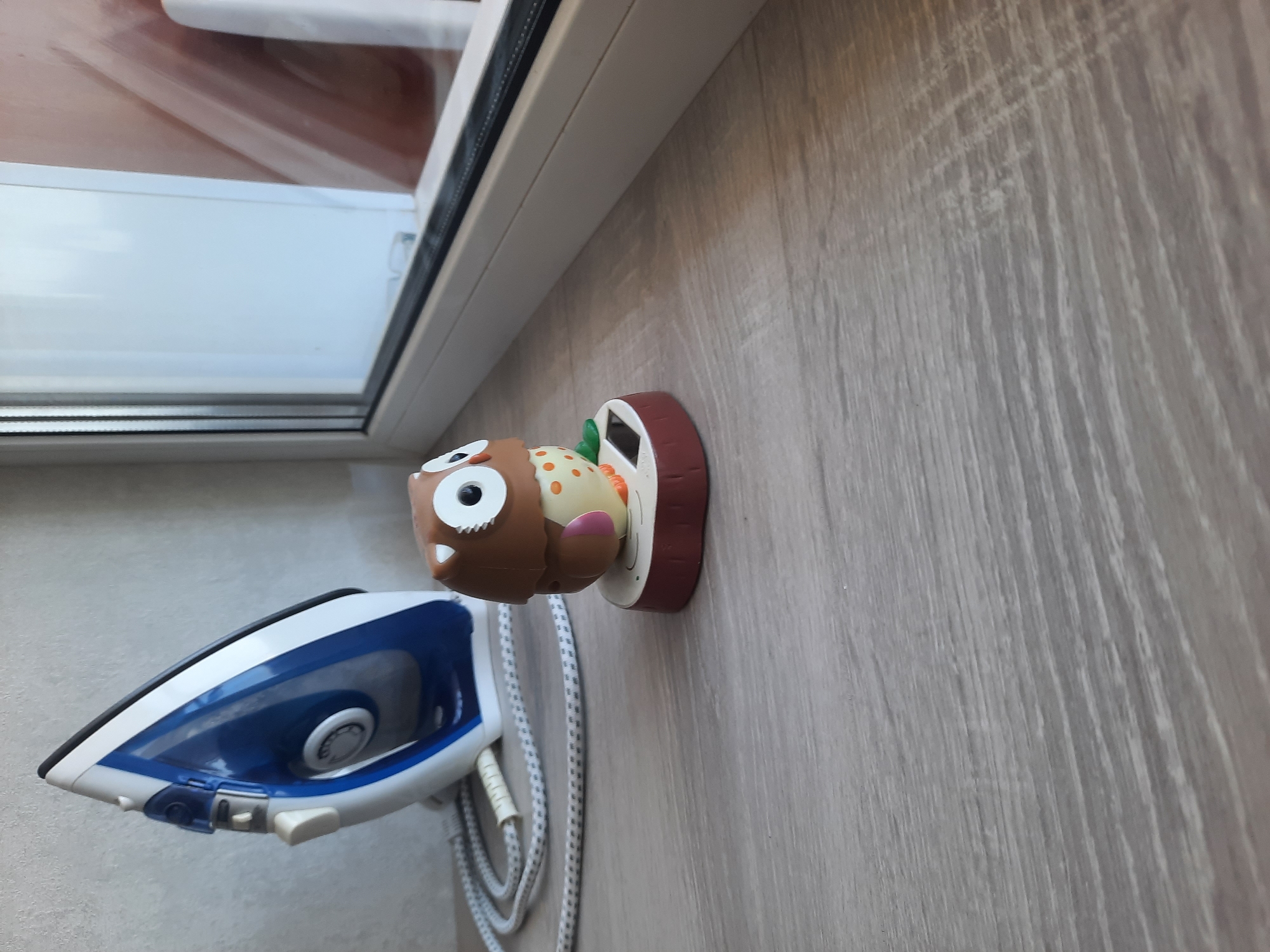 An owl figurine with a solar-powered waving mechanism sits on a wooden surface next to an iron and a window. The owl is brown and white with orange accents and large, round eyes. It stands on a red base that resembles a tree stump, with a small solar panel visible on the front. To the left of the owl is a blue and white iron with its cord coiled nearby. The scene is illuminated by natural light coming from the window, which has a white frame and a reflective surface. The wooden surface appears to be a countertop or shelf. The background is a light-colored wall.