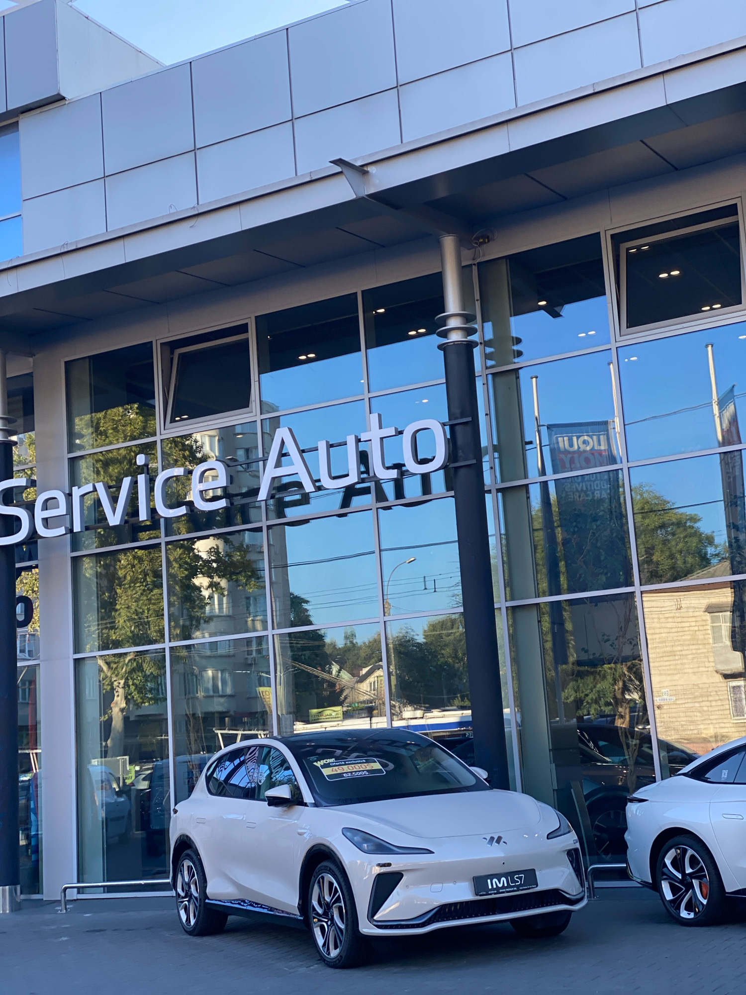 The image shows a white electric car, identified as an "IM L57", parked in front of a modern building with large glass windows. The building displays a sign that reads "Service Auto". Reflections in the windows show trees, buildings, and a banner for "LIQUI MOLY". Another white car is partially visible to the right. The overall scene suggests a car dealership or service center in an urban environment. Given the presence of the "IM L57" car, which is associated with the IM Motors brand, and the context of automotive services, this is likely a dealership in Chișinău, Moldova, as indicated by external information. The sky is blue with some clouds, and the lighting suggests it is daytime, likely late afternoon or early evening due to the angle of the sun. There are no people visible in the foreground or midground of the image.