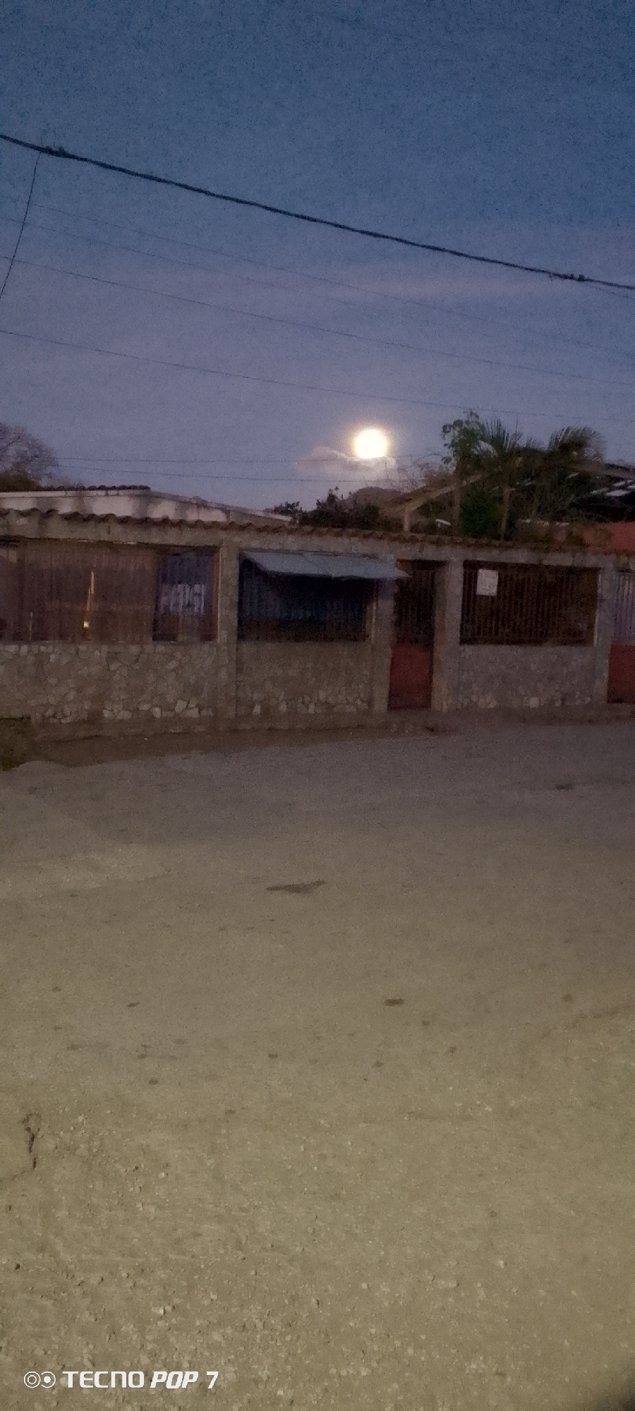 The image depicts a street scene in Altagracia, Venezuela at twilight or night. The sky is a deep blue, with the moon visible above a low-lying building. The building is a simple, one-story structure with a mix of stone and metal siding, and what appears to be advertisements or signage on the facade. There is a dirt or paved road in front of the building. Power lines stretch across the top portion of the image. No people are visible. The overall environment appears rural or underdeveloped.
