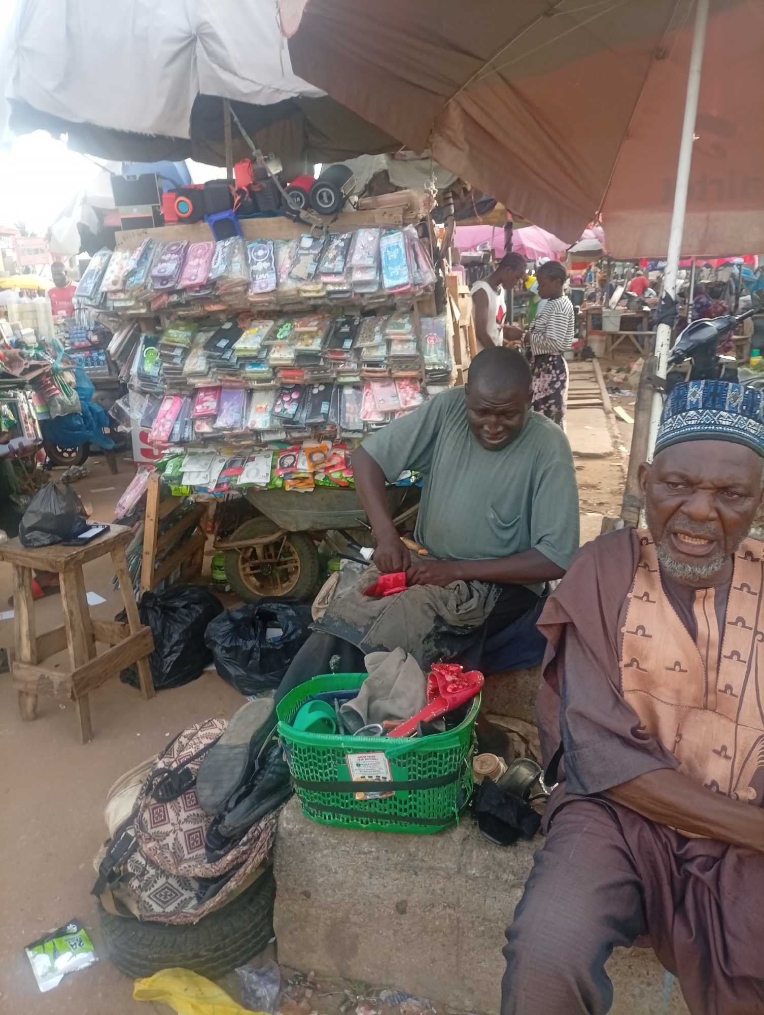 The photo depicts an outdoor scene in Sabon Tasha, Nigeria. An older man wearing a traditional patterned gown and a blue patterned cap sits on a concrete block, looking towards the camera. Next to him, a younger man in a green shirt sits and appears to be working on a red object with a tool. In front of them is a green plastic basket containing various items, including a red shoe. Behind the men, a large display of phone cases is arranged on a wooden structure. Other market goers and stalls are visible in the background, along with a parked moped, all under the shade of various umbrellas. A small wooden stool sits to the left with a bag on it. Black plastic bags are scattered around the area.