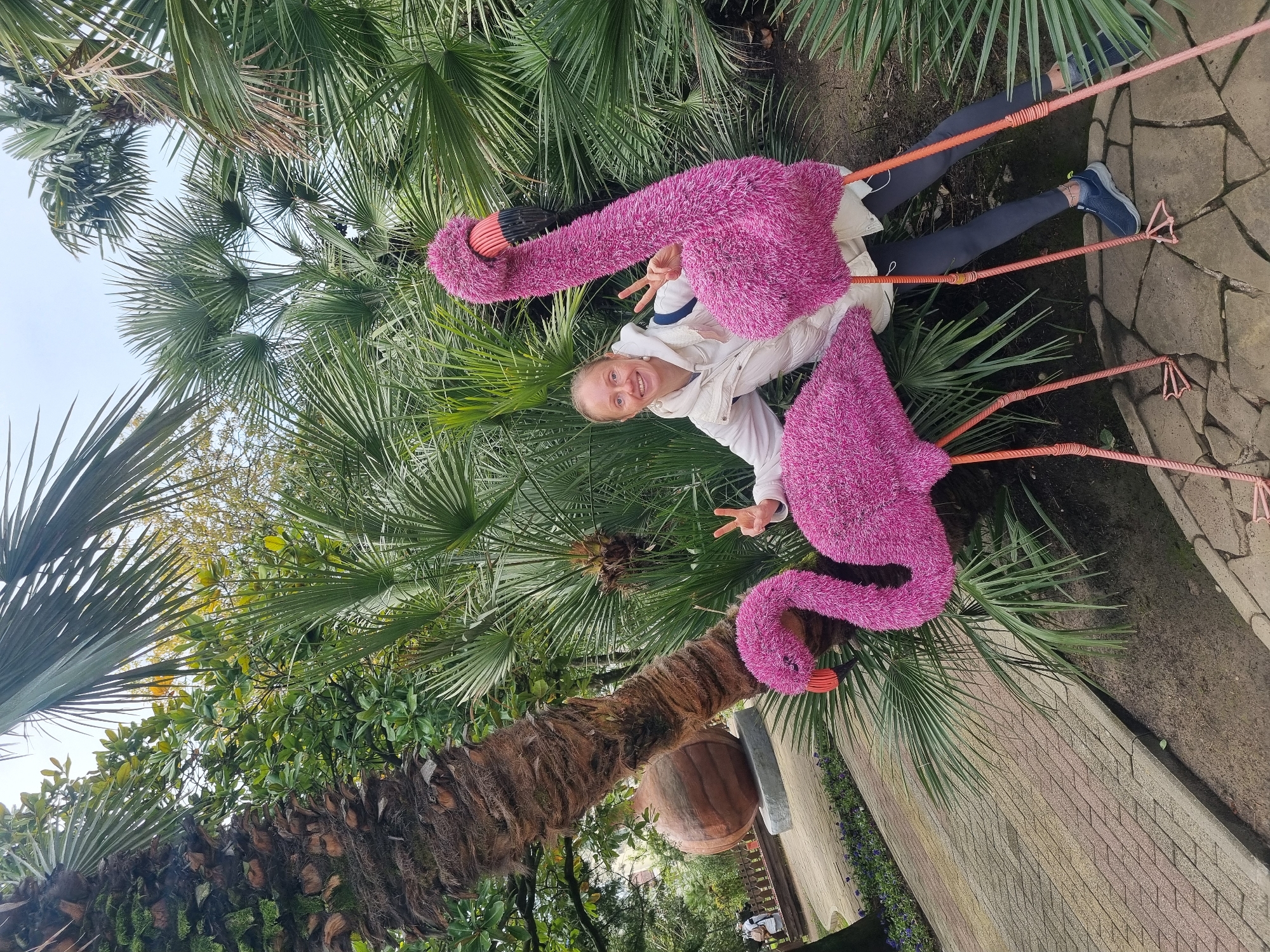 A woman poses with two pink flamingo props in Sochi, Russia. She lies on her back amidst lush vegetation, including palm trees, and a stone-paved path. The woman wears a white jacket, dark leggings, and blue sneakers. She holds up two fingers in a peace sign with each hand. The flamingo props have long, pink legs and pink, fuzzy bodies. One flamingo's head is black and red. A brown tree trunk and a large, brown pot are visible in the background.