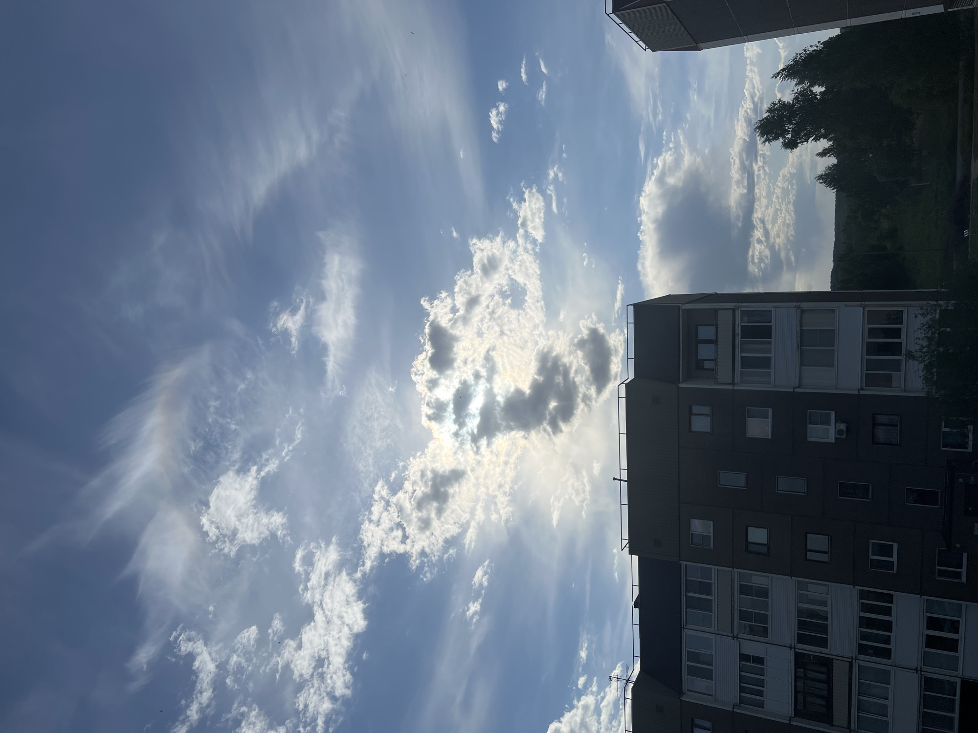 The image, taken in Марковский, Russia, presents a view of the sky with clouds, buildings, and vegetation. The sky is predominantly blue with streaks of white cirrus clouds and a cluster of puffy cumulus clouds partially obscuring the sun. The sunlight streams through the gaps in the clouds. Two multi-story apartment buildings frame the sides of the image. One building has multiple rows of windows and is likely residential. Vegetation, possibly trees, can be seen in the background. The overall scene is a daytime cityscape, focusing on the interplay of clouds, sky, and urban architecture.