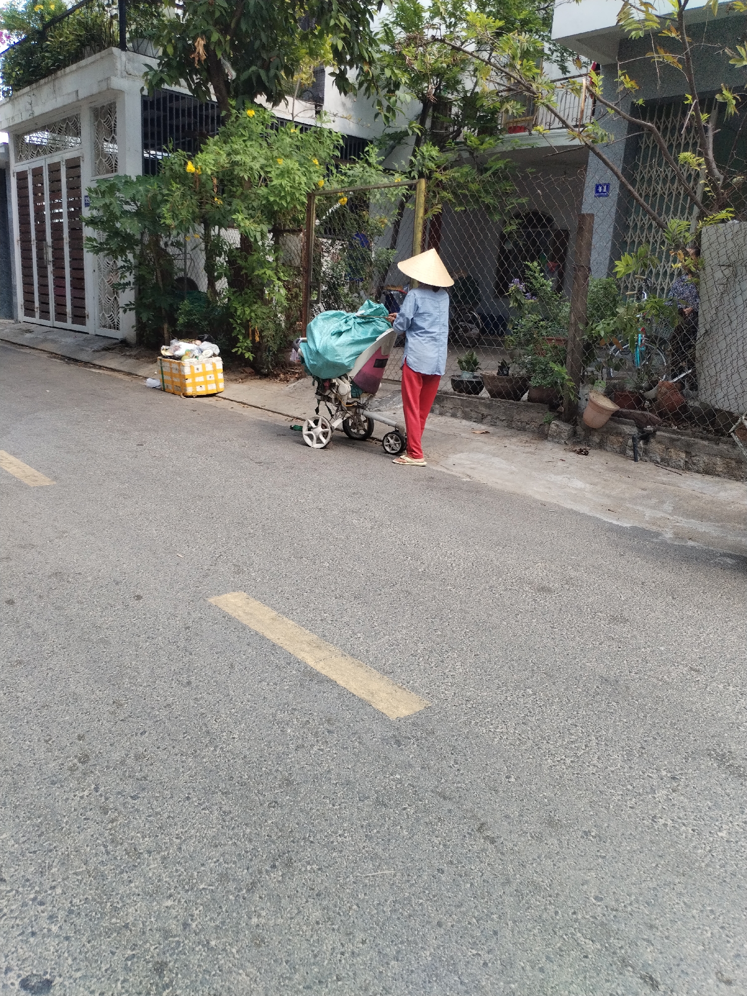 The image shows a woman in Bac Nha Trang Ward, Vietnam, wearing a traditional conical hat, a light blue shirt, and red pants, pushing a small cart filled with a large green bag. The cart also has a pink object on it. Next to the cart is a large yellow and white box. The woman is on a paved street with yellow road markings. Behind the woman is a fence and a house with a gray exterior. There are plants and trees around the fence. Another house is visible in the background on the left with a white and brown gate.