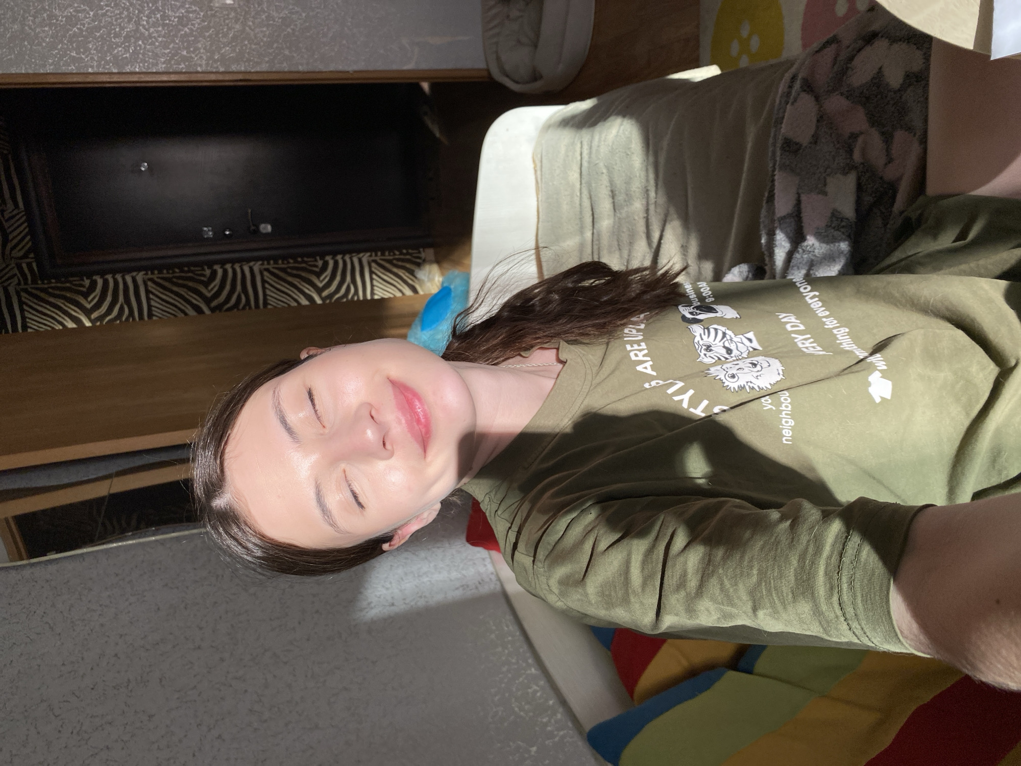 A person with closed eyes is lying on a couch indoors. The individual has dark hair and is wearing an olive green long-sleeved shirt. The background includes a dark television screen above a patterned surface, a partially visible cream-colored object that could be a cushion or pillow, and a corner of what appears to be a colorful striped pillow or blanket. The image is brightly lit, and shadows are cast across the person's face and clothing. The location and city are not discernible from the image alone.