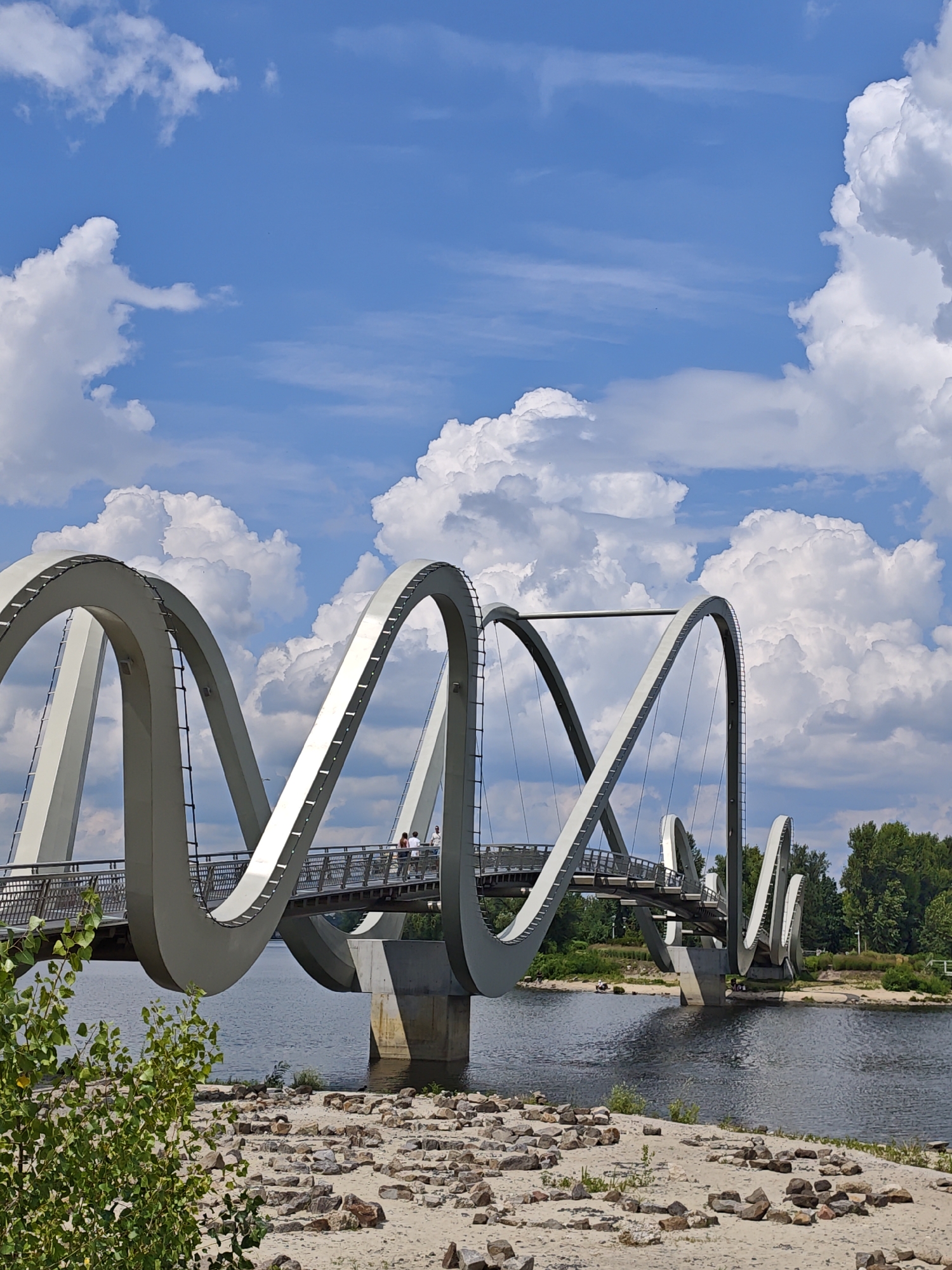 The image shows the People's Friendship Arch in Kyiv, Ukraine, a monument with a distinctive ribbon-like design. The arch is made of grey steel and spans over a body of water, likely the Dnieper River. The sky above is a bright blue with scattered white, fluffy clouds. The foreground consists of a sandy riverbank with numerous stones. A bridge, part of the arch structure, is visible with a few people walking on it. Lush green trees are seen on the far bank of the river. The overall scene depicts a sunny day at a prominent landmark in Kyiv.