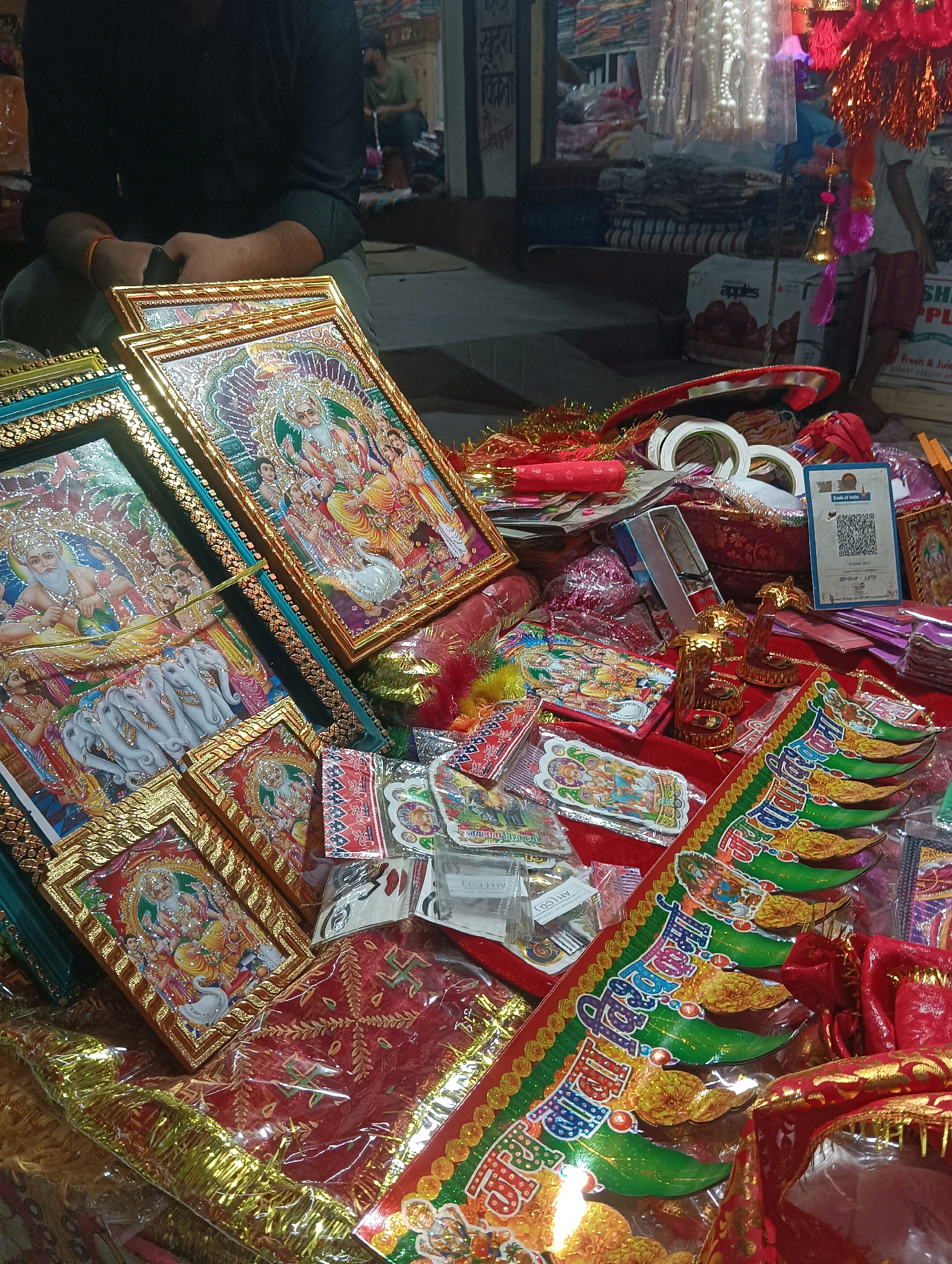 In the city of Dakhin Parsa, India, a stall vendor, a male wearing a dark long-sleeved shirt and grey pants, sits cross-legged amidst a vibrant display of religious artifacts and decorative items. His arms are crossed, and a thin orange band is visible on his left wrist. The stall is filled with numerous framed pictures of deities, predominantly featuring Hindu gods and goddesses in ornate, golden frames. Among these are images of a bearded male deity with multiple arms, a deity with an elephant head, and a depiction of several white elephants.
The foreground is dominated by these framed pictures, along with various decorative elements like red fabric adorned with gold embroidery, shimmering gold tassels, and colorful paper cutouts. Several rolls of white tape are also visible. In the background, shelves are stacked with more merchandise, including what appear to be fabric rolls and packaged goods. Two other individuals are partially visible in the background; one is a male seated in the distance, and another, possibly a child, is standing. The overall scene suggests a bustling marketplace during a festive season, with an emphasis on religious and celebratory items. A QR code for "Bank of India" is prominently displayed on a light blue card, indicating a modern transactional element within the traditional setting.