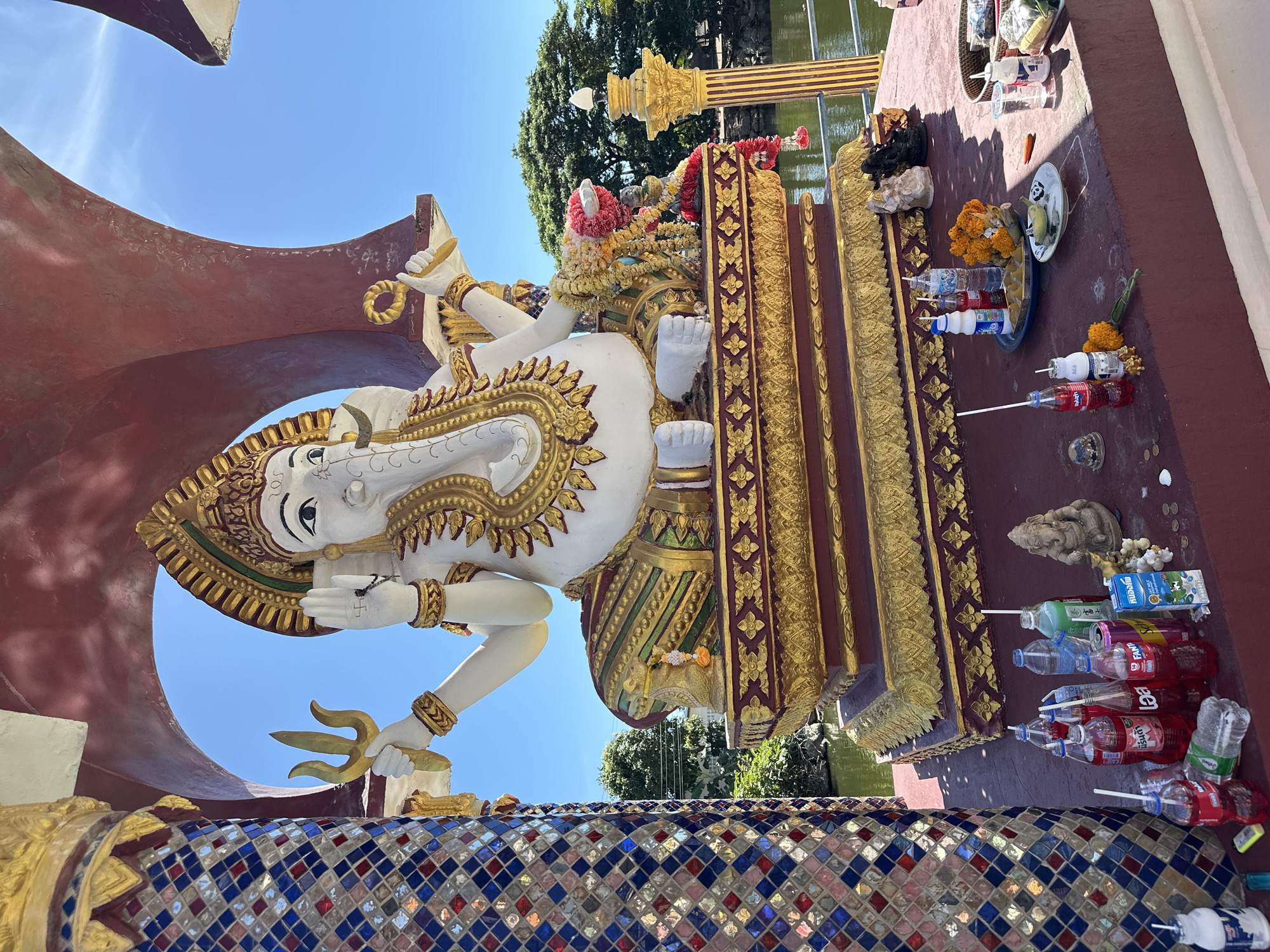In Baan Plai Laem, Thailand, a large, ornate statue of the deity Ganesha sits prominently on a raised platform. The statue is predominantly white with intricate gold and green detailing, adorned with a garland of colorful flowers. Ganesha is depicted seated, with multiple arms, holding various symbolic objects. The platform itself is richly decorated with gold trim and red accents.
Surrounding the statue, on the platform and at its base, are offerings, likely left by devotees. These include numerous plastic bottles of drinks (some with straws), small figurines of deities, coins, and plates of food, primarily consisting of marigold flowers and what appears to be fruit. A water-filled area, possibly a pond or canal, is visible in the background, bordered by trees and a clear blue sky. The scene is framed by decorative pillars, one of which is covered in colorful, mosaic-like tiles, and a red, arch-like structure above the statue. The lighting suggests it is a bright, sunny day.