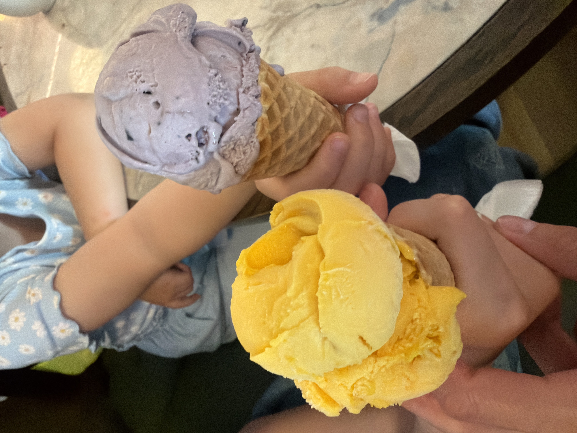 This image displays an indoor scene featuring two individuals holding ice cream cones. While the requested location is Dalaman, Turkey, no specific visual identifiers for this city are present in the image. In the immediate foreground, an adult hand holds a waffle cone containing a large scoop of bright yellow ice cream, with white napkin material partially visible beneath. Behind this, a child, wearing a light blue top with white daisy patterns and light blue lower garments, holds a separate waffle cone with a large scoop of light purple ice cream. The child's arms are positioned with one arm partially crossed over the other, holding the cone in the right hand. The environment includes a light-colored, possibly marble-patterned tabletop visible on the right, and a dark, likely upholstered, seating surface in the left background. The ice cream in both cones exhibits minor textural variations consistent with light melting.