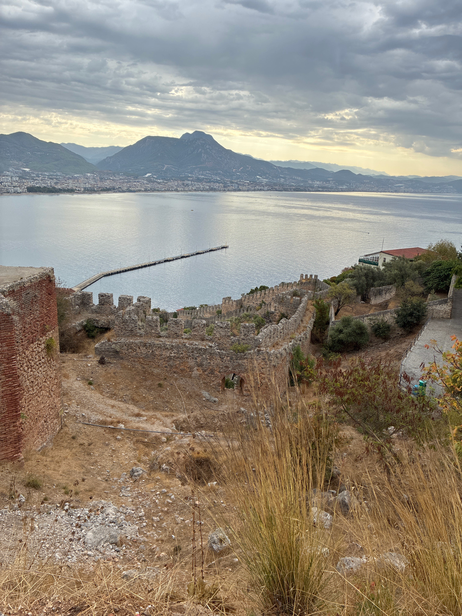 This is a medium-shot, eye-level, vertical photograph taken from an elevated vantage point overlooking the city of Alanya, Turkey. In the foreground, the weathered stone ruins of an ancient fortress wall and a brick structure are visible, with dry, tall grasses growing in front of them. The middle ground shows the continuation of the stone fortress walls winding along the hillside, with trees and other vegetation interspersed. Beyond the fortress, a calm, grey sea stretches to the horizon. A long pier extends from the city into the water. The background features a densely populated coastal city with buildings nestled against a backdrop of rolling, hazy mountains under a cloudy sky. The overall impression is one of historical significance and a panoramic view of the coastal landscape.