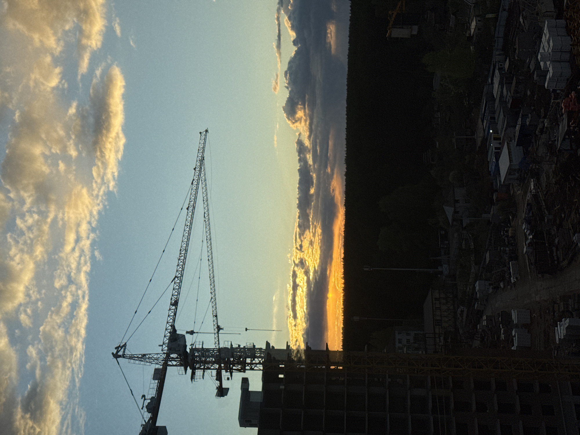 The image shows a construction site in Izhevsk, Russia, during sunset. A tall crane stands next to an unfinished concrete building. The sky is blue with golden clouds on the left side. On the right, darker clouds line the horizon where the sun is setting, casting a golden glow. In the background, a dark forest is visible, bordering the construction area. Construction materials and equipment are scattered on the ground around the building.