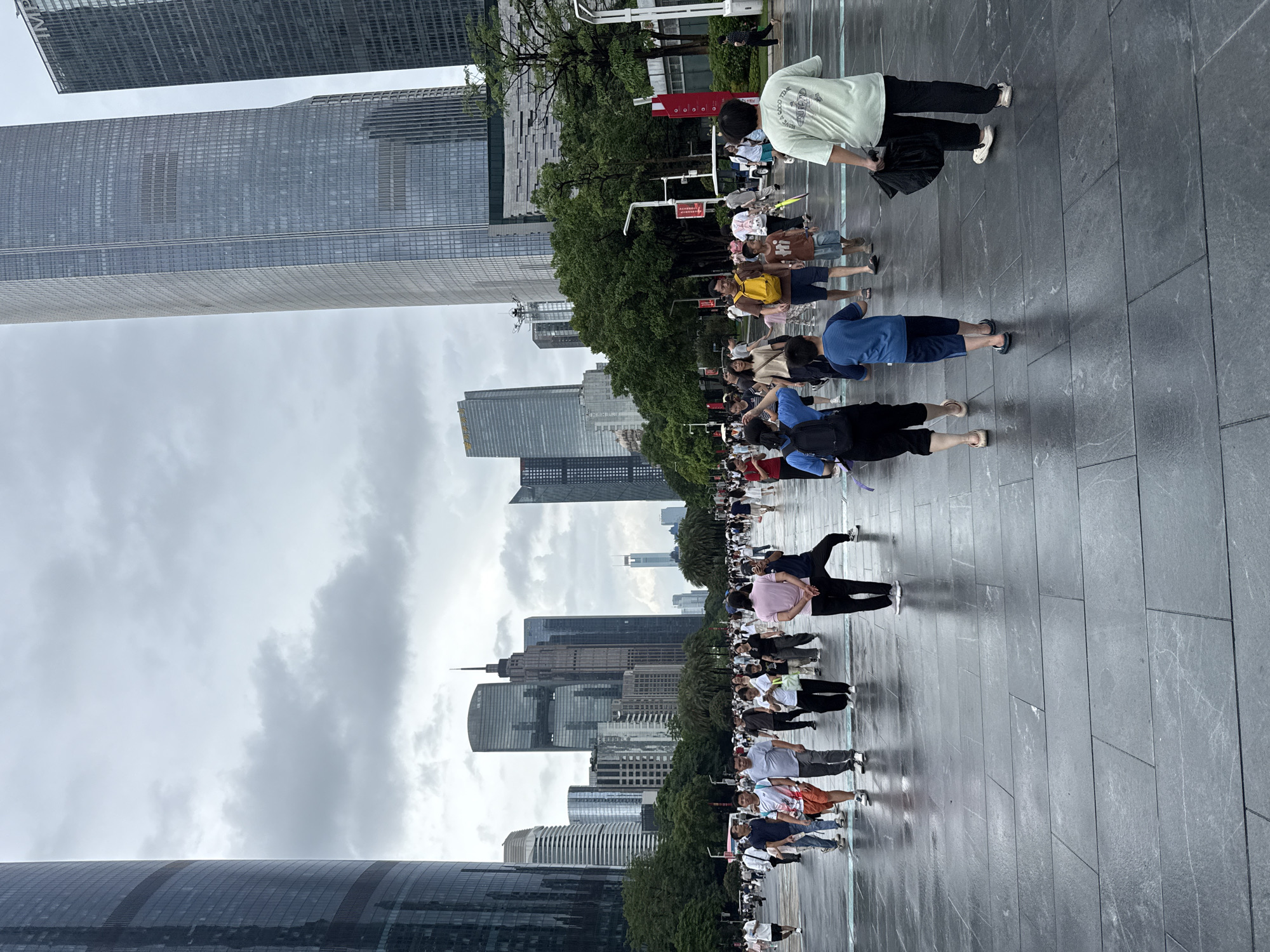 The image depicts a wide, paved plaza in Tianhe District, China, with numerous people walking. Towering modern skyscrapers with glass facades dominate the background under a cloudy sky. The paving stones are wet, suggesting recent rain, and reflect the surrounding buildings and sky. People are dressed in casual summer attire, and many are walking in various directions across the plaza. Some individuals are walking alone, while others are in groups or families. The scene is populated by a diverse range of ages, including children and adults. A few trees and green spaces are visible interspersed between the buildings, providing a contrast to the urban architecture. The overall impression is that of a busy public space within a modern, developed city.