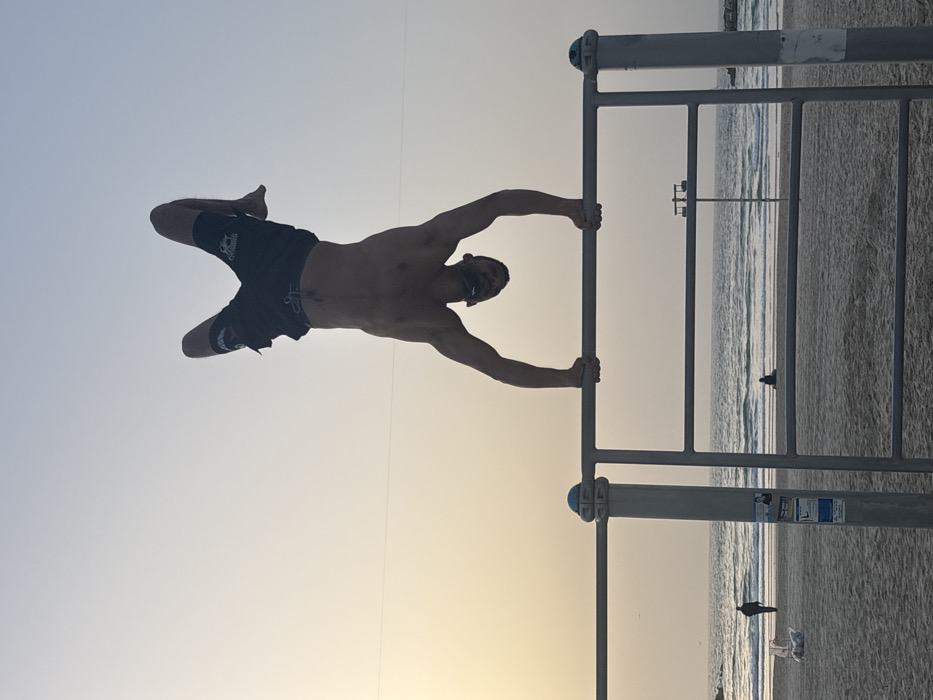 A muscular man is performing a human flag on a metal bar structure, silhouetted against a light sky and beach backdrop. He's holding the bar with straight arms, his body extended horizontally. His legs are slightly bent and positioned upwards. The bar is set up on a beach near the ocean, visible through the metal frame along with a few scattered people and birds.
