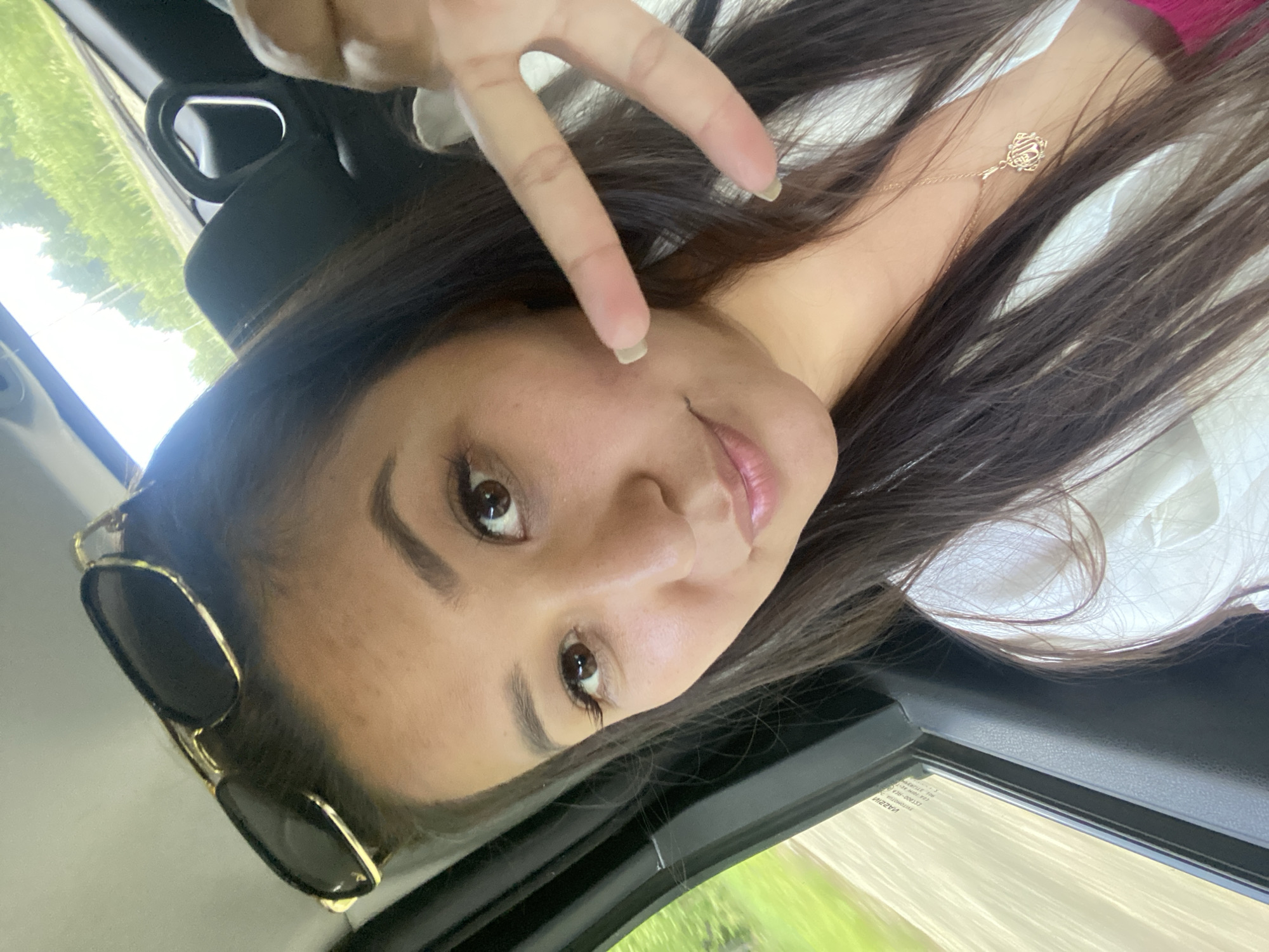 A woman is taking a selfie from inside a vehicle. She is wearing sunglasses on her head and a white shirt with a pink top underneath. A gold pendant necklace is visible on her neck. Her right hand is raised, with two fingers extended, making a peace sign. The background shows greenery and trees, suggesting a rural or suburban setting. The image does not provide any information about the city of Zilair, Russia.