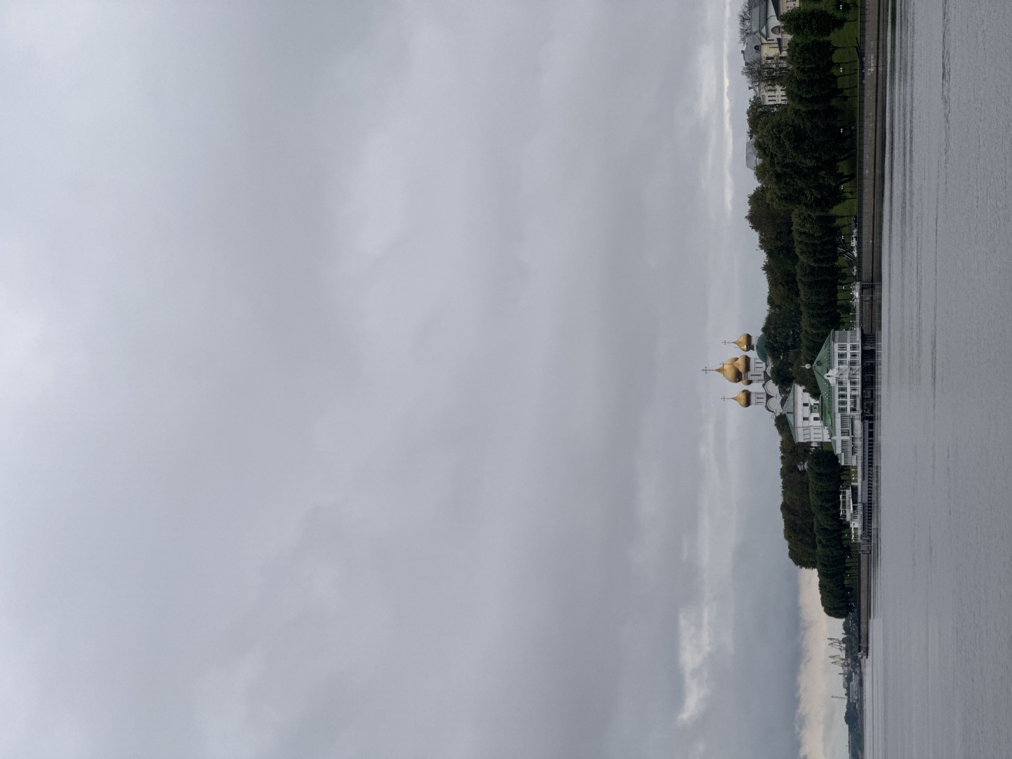 The image shows a waterfront scene with a cloudy sky. In the foreground, a body of water reflects the overcast sky. Along the embankment, a line of trees separates the water from a low-rise building complex. To the left of the building complex, a prominent church with golden domes is visible on a hill. Further in the distance, industrial cranes can be seen along the horizon. The scene is devoid of any people. The location is identified as Shebunino, Russia.