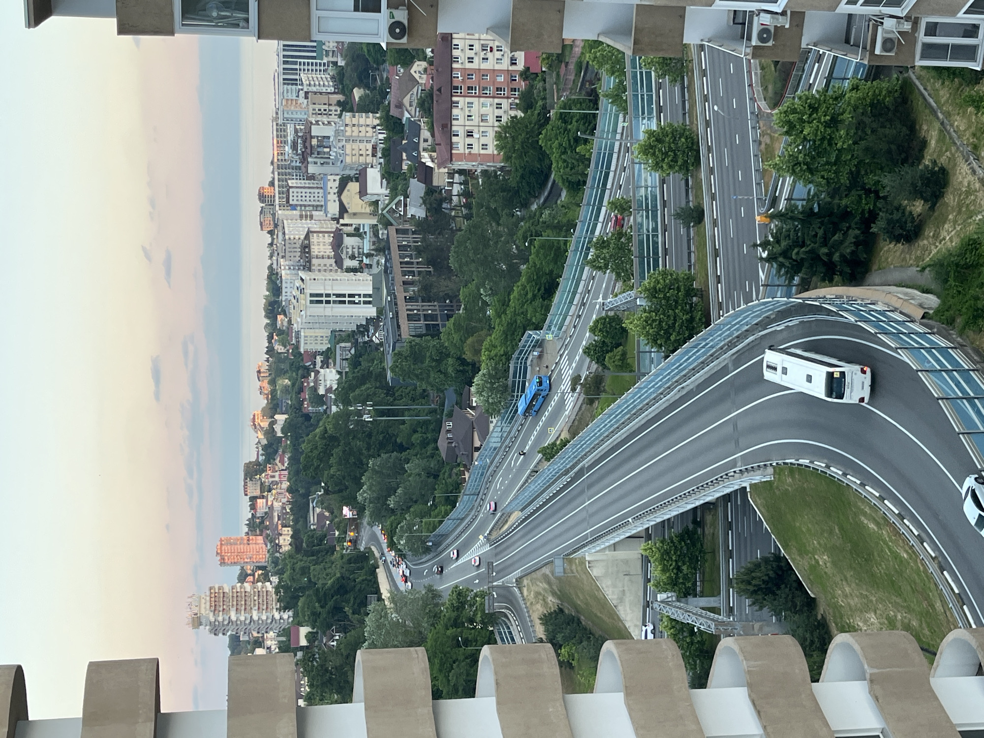 This is a high-angle, eye-level shot of a multi-level highway intersection in Sochi, Russia, with buildings and the sea in the background. The sky is a soft pink and blue, suggesting either sunrise or sunset. On the left and right sides of the frame, parts of a modern apartment building with curved balconies are visible, framing the city view.
The highway is paved with asphalt and marked with white lines. Several cars are driving on the highway. A blue bus is visible on the upper level of the highway, and a white minibus is on the lower level. The highway is bordered by protective barriers.
The city itself is a mix of modern high-rise buildings and older, smaller structures, interspersed with lush green trees. In the distance, the Black Sea is visible, with a hazy horizon. The overall impression is of a developed urban environment with significant infrastructure, set against a natural backdrop.