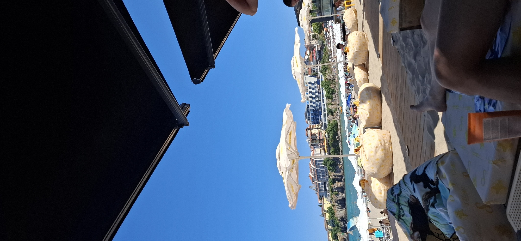 The image depicts a sunny day at a beach in Sozopol, Bulgaria. In the foreground, several large, light-colored beanbag chairs are arranged on the sand, some adorned with star patterns. A person's legs and feet are visible, relaxed on a beach lounger. A sunblock bottle sits on the lounger. In the middle ground, several white beach umbrellas provide shade. Beyond the umbrellas, the turquoise water of the Black Sea is visible, with some people swimming. The background features a coastal town with buildings situated on a cliffside. The sky is a clear, vibrant blue. The overall scene conveys a relaxed and leisurely atmosphere at a popular tourist destination.