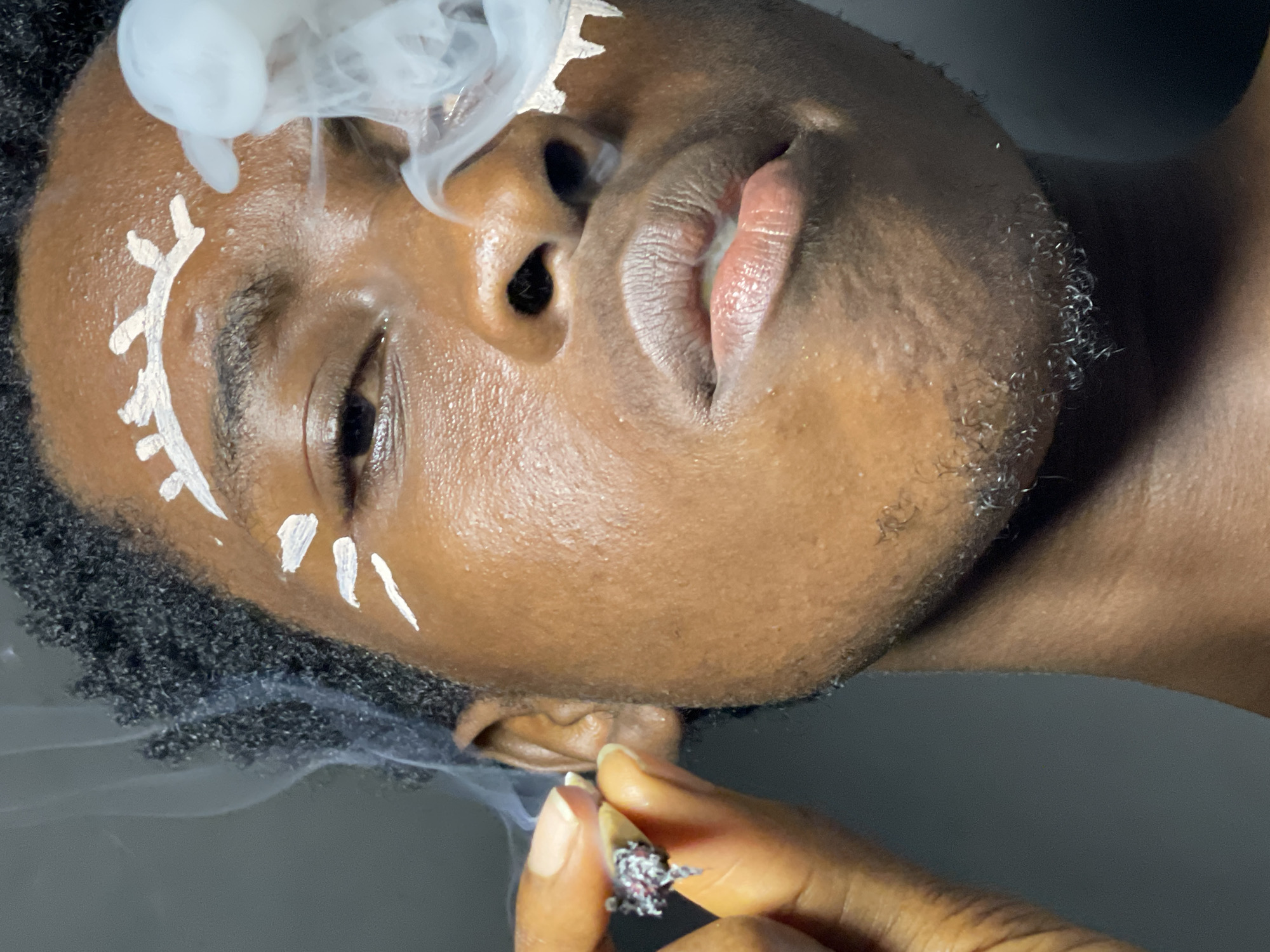 A close-up shot features a person with dark skin and short, curly black hair. White face paint adorns their forehead and cheeks, with markings resembling stylized eyebrows and stripes on the cheeks. The person's right hand, with manicured nails, holds a lit cigarette, from which smoke billows around their face. The background is a plain, dark gray surface. The scene does not provide enough information to determine a specific location within Nkpor, Nigeria.
