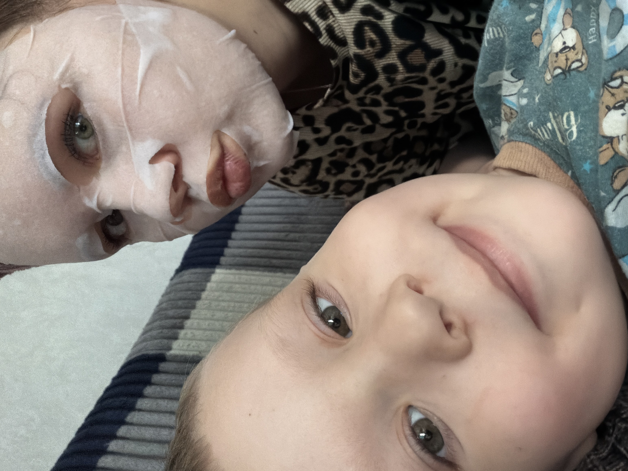 An overhead close-up image captures an adult and a child lying down, looking upwards. The adult, positioned on the left, has light skin and striking green eyes, which are visible through the eye-holes of a white sheet face mask covering their face. Dark eyelashes frame their eyes, and their lips are slightly parted through the mask's mouth opening. They are wearing a garment or blanket with a distinct brown and black leopard print pattern.
To the right of the adult, a young child with light skin and light brown or greenish eyes is also lying down, facing the camera with a gentle smile. Their light-colored hair is visible at the top of their head. The child is dressed in a blue-grey long-sleeved top or pajamas adorned with cartoon-like bear or animal figures and faint white text, including partially discernible words like "Happy" and "Surly."
Both individuals appear relaxed, suggesting a casual, intimate moment, possibly at home. The adult is engaged in a skincare routine with the face mask. A striped fabric, featuring shades of grey, navy blue, and white, is visible in the background, likely part of bedding or a blanket. The lighting is soft and even, indicating an indoor setting. The image conveys a sense of quiet bonding or a shared, relaxed activity.