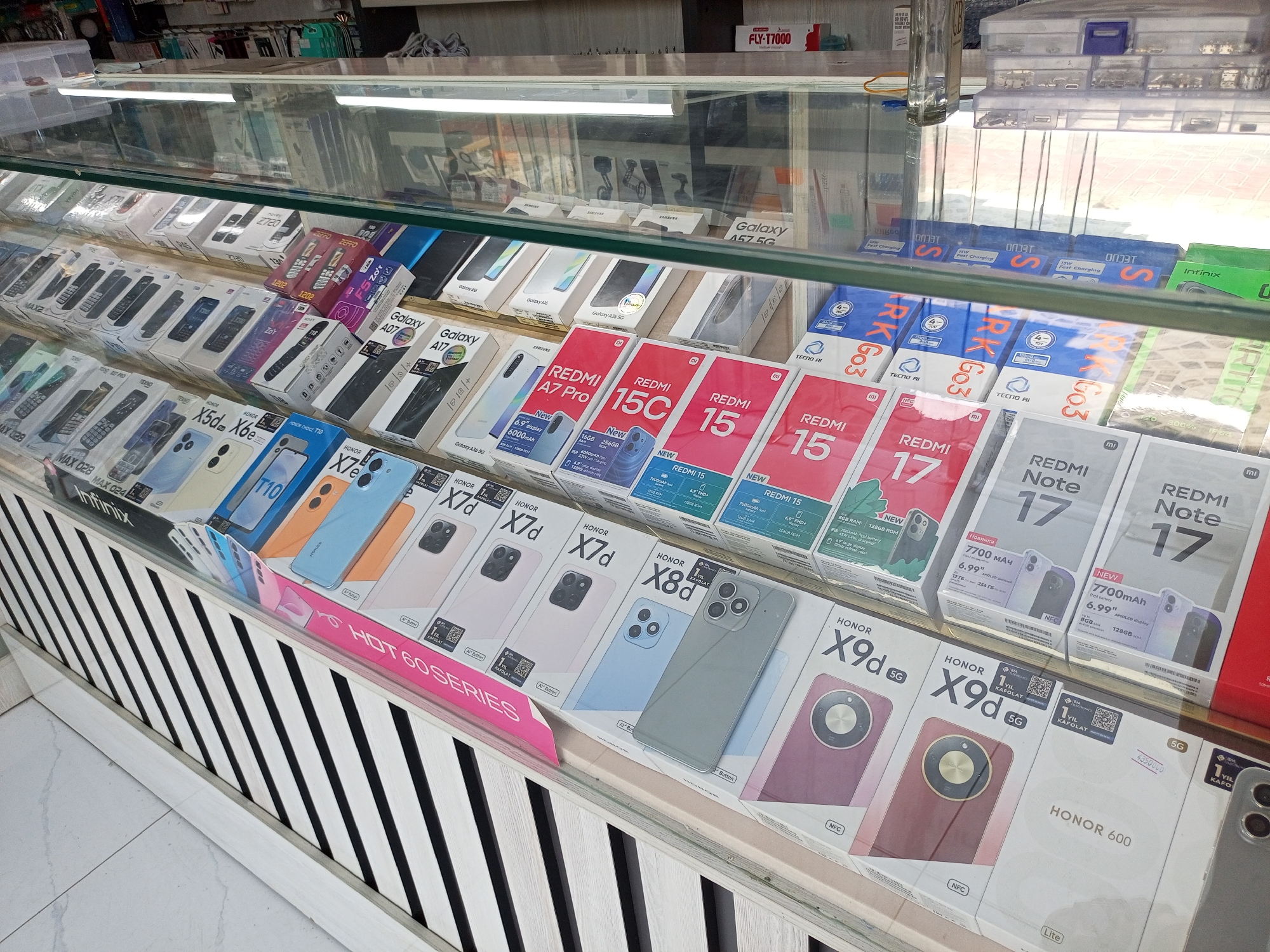 The image shows a retail display of mobile phones inside a glass-enclosed cabinet at a store in Shahrisabz, Uzbekistan. The phones are arranged in rows, mostly in their original packaging. Various brands and models are visible, including Samsung Galaxy A series, Redmi, Honor, and Tecno. Some packages display specifications such as screen size, battery capacity, RAM, and storage. The display also features promotional banners and pricing information. The background suggests a commercial environment with other retail goods and shelving.