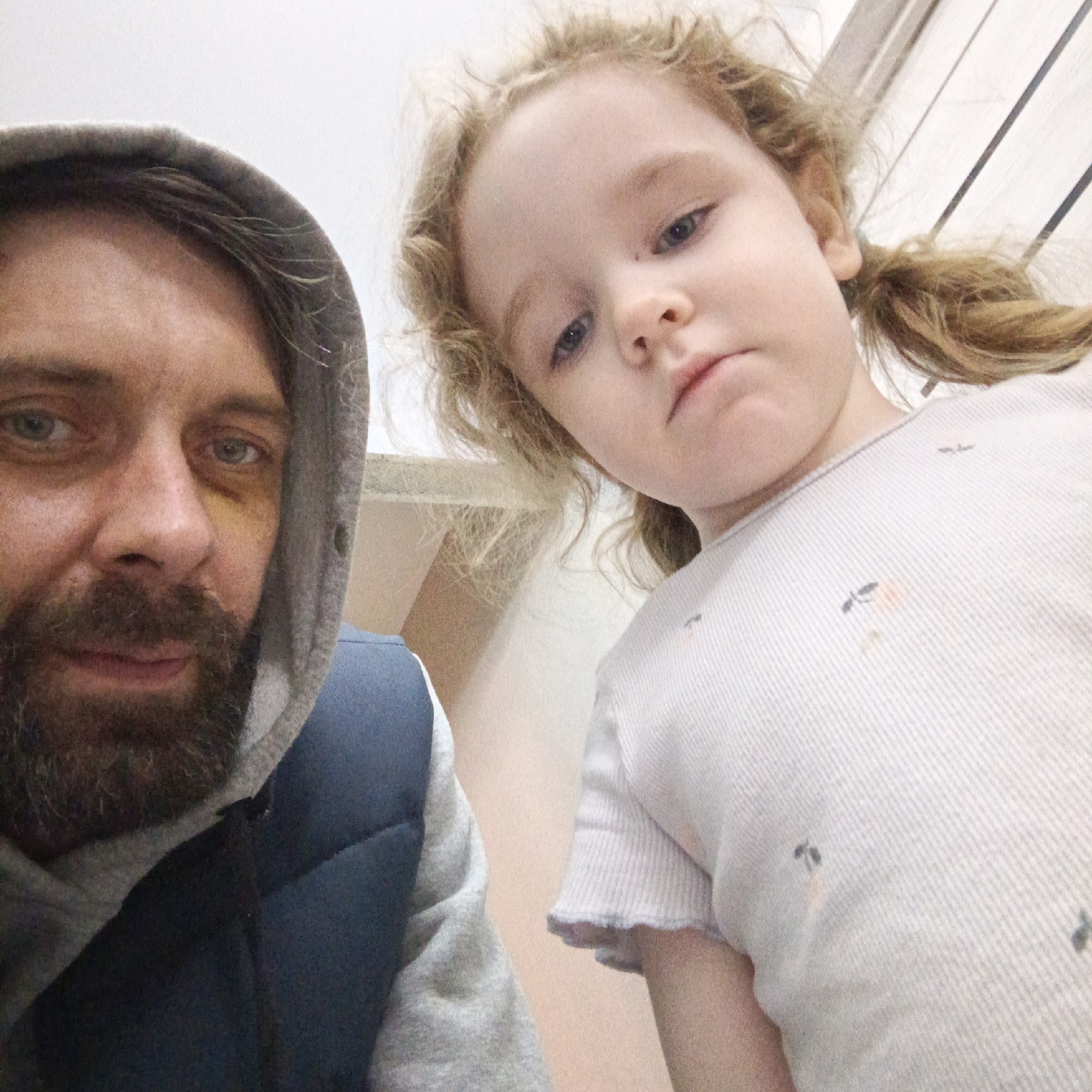 A low-angle, close-up selfie captures a man and a young child indoors. On the left, a man with a beard and dark hair wears a grey hooded sweatshirt and a blue puffer vest. He has blue-green eyes and a noticeable black eye or bruise under his right eye, giving him a neutral to slightly somber expression as he gazes at the camera.
To his right, a young child, likely a girl, looks directly into the camera with a serious or pensive look. She has curly blonde or light brown hair styled into two small pigtails. Her eyes are blue. She is dressed in a light-colored, possibly white or light grey, ribbed top featuring small, dark, abstract patterns.
The background is predominantly plain and light-colored, with faint vertical lines visible in the upper right quadrant, suggesting an interior wall or ceiling. The informal attire and indoor setting point to a casual environment, potentially a home in Марковский, Russia. No text is visible within the image.