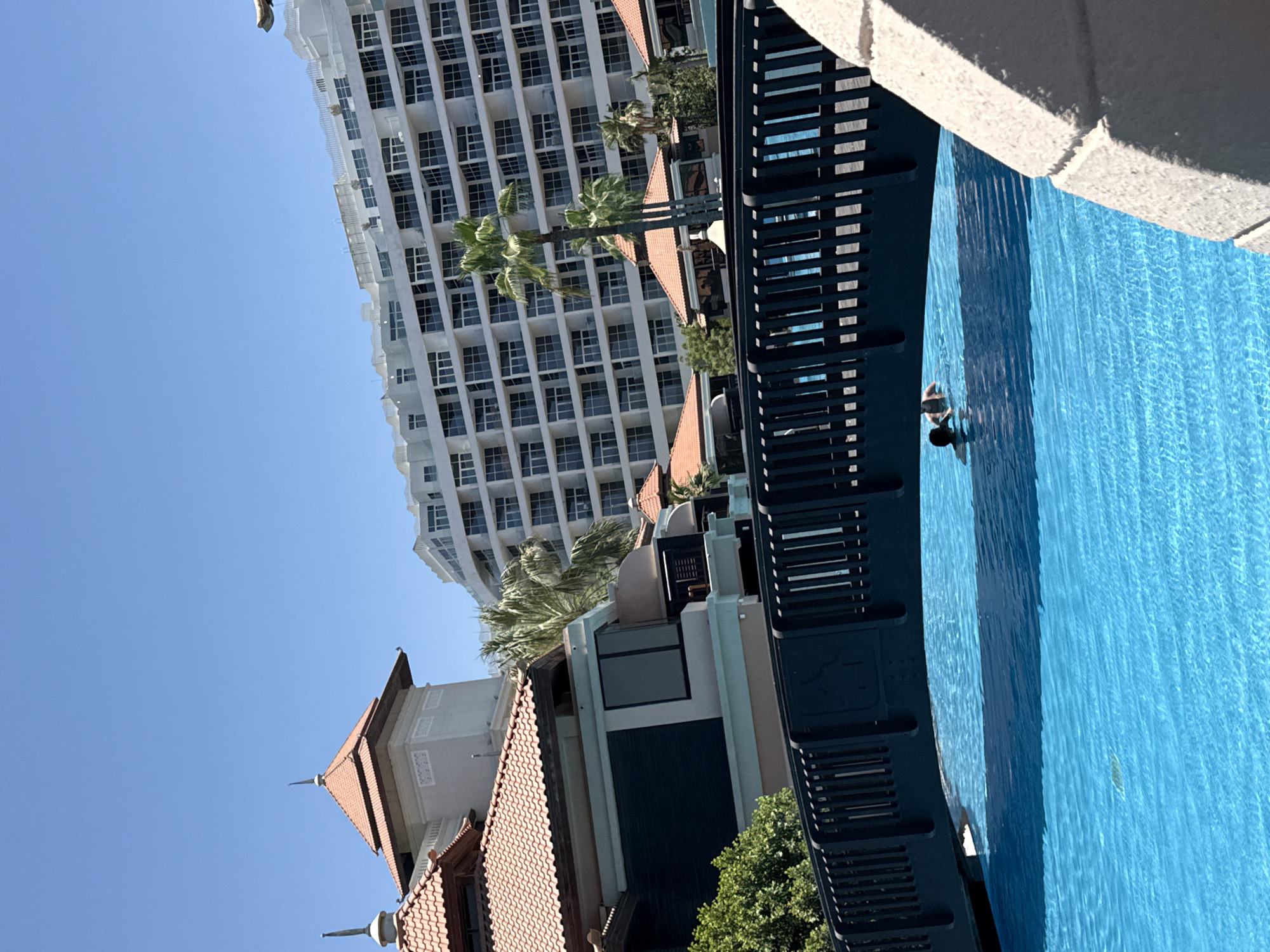 The image depicts a sunny day at a resort in Dubai, United Arab Emirates. In the foreground, a bright blue swimming pool fills the lower half of the frame, with two individuals visible swimming near a dark-railed bridge that arches over the water. The bridge casts a shadow on the pool's surface. Behind the pool, a section of a resort building with terracotta tiled roofs and light-colored walls is visible, with lush green foliage interspersed. Further in the background, a large, modern white hotel building with numerous balconies and windows dominates the skyline under a clear blue sky. The architecture suggests a luxurious vacation destination.