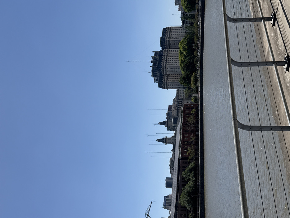 A cityscape is viewed from a high vantage point along a waterfront. Tall buildings line the shore, with some featuring intricate spires and towers. A crane is visible in the lower portion of the frame. The water stretches out to meet the buildings. A metal railing and a weathered wooden deck are in the foreground. The sky is a clear, vibrant blue.