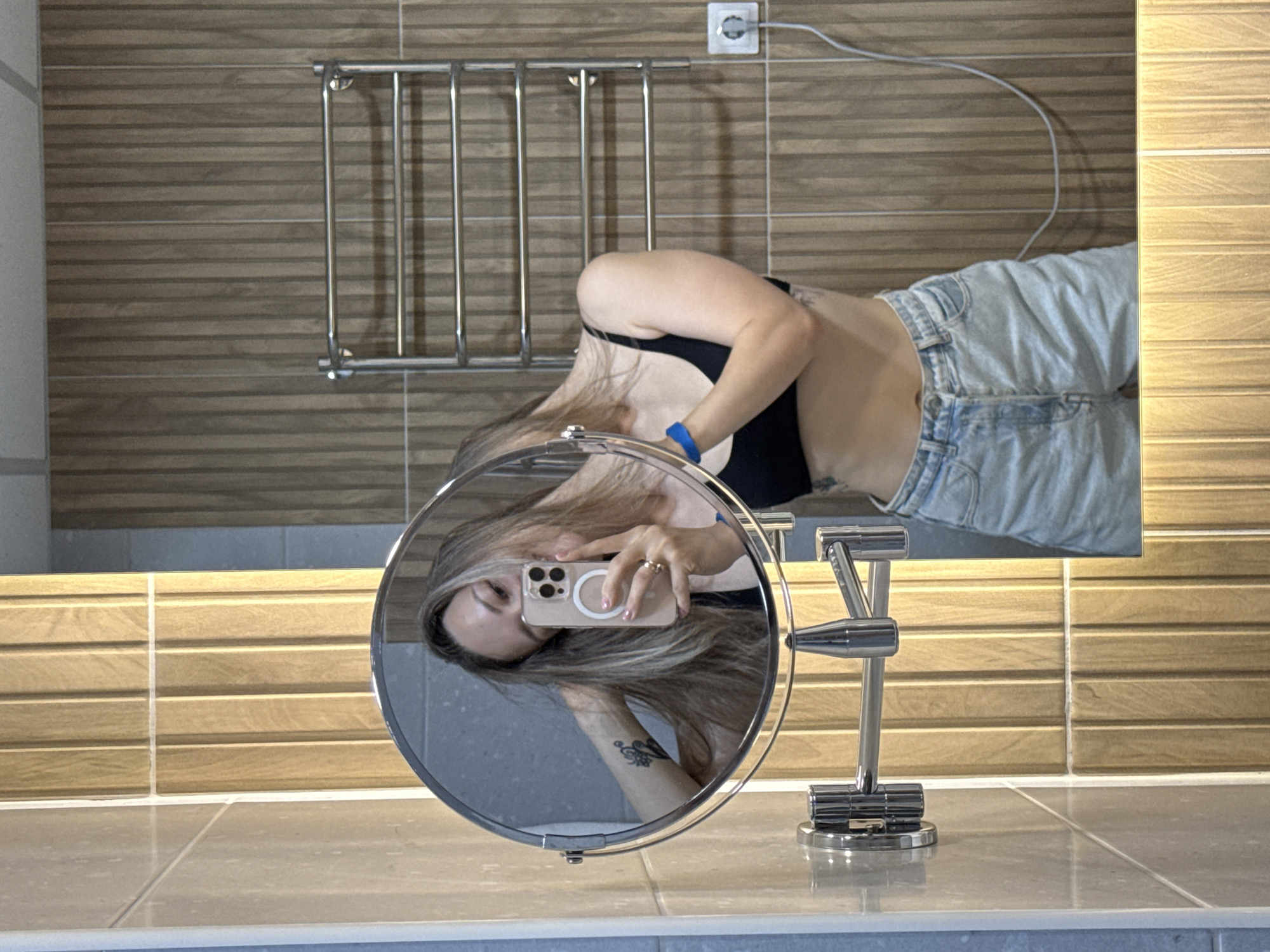 A female subject is depicted in a bathroom selfie taken in Nebug, Russia. She has long, light-colored hair and is wearing a black sports bra and light-wash denim shorts. A tattoo featuring a floral or abstract design is visible on her left upper arm. A blue wristband is present on her right wrist, and a ring is on her left ring finger. She holds a light-colored smartphone in her left hand, which is visible in an adjustable circular magnifying mirror. Her right hand is positioned in her hair. The magnifying mirror, mounted on a chrome articulated arm, is itself reflected in a larger wall mirror. The bathroom walls are tiled with vertical wood-grain patterns in varying shades, appearing light beige on the left and a darker brown behind the towel rack. A chrome towel rack is mounted on the wall to the subject's right. A white power outlet with a white charging cable plugged in is visible below the towel rack.
