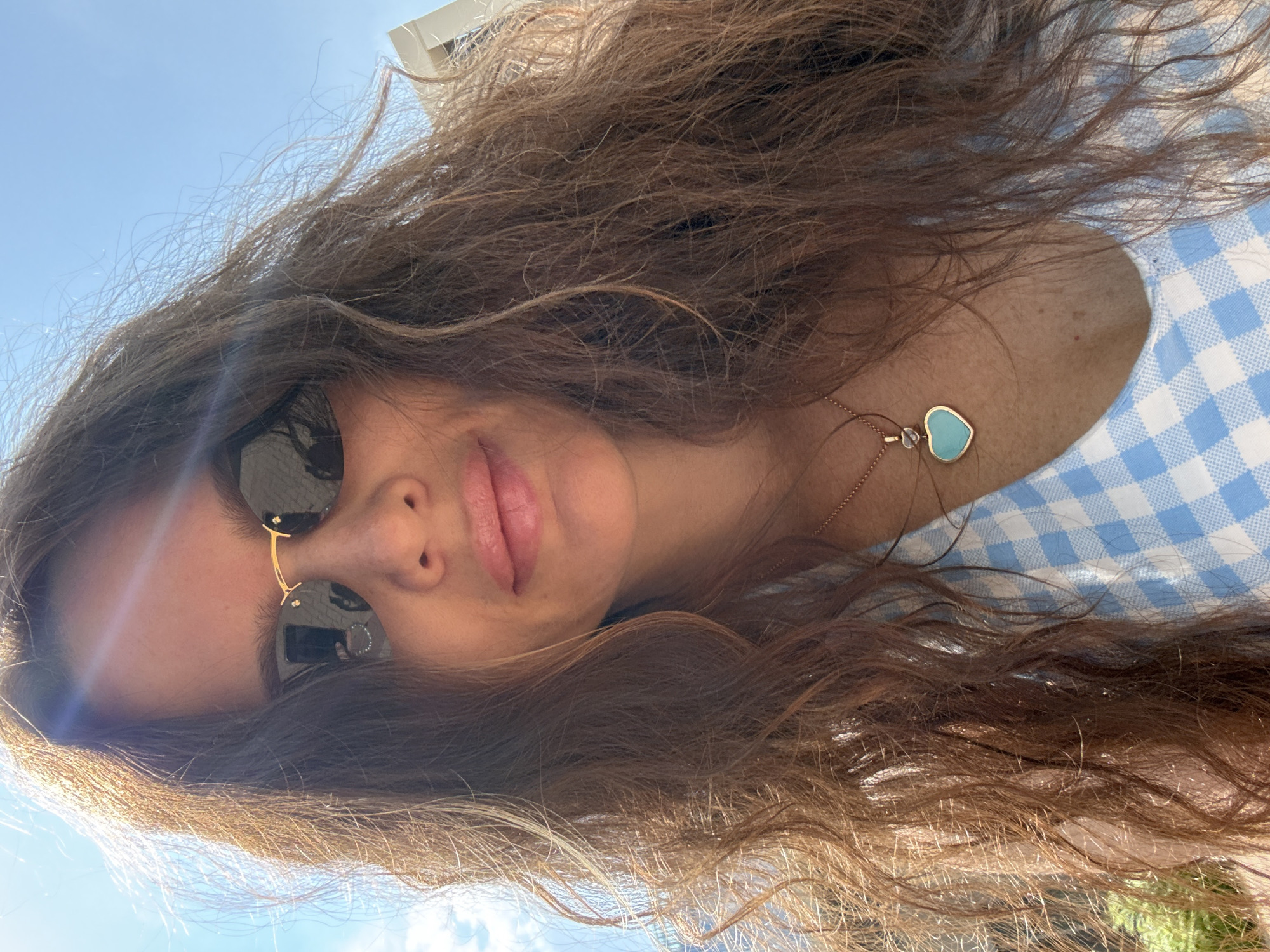 A woman with long, wavy brown hair and sunglasses is taking a selfie outdoors on a sunny day. She is wearing a blue and white checkered top and a necklace with a turquoise heart-shaped pendant. The background shows a bright blue sky and some greenery. The image does not provide any information about the location in Moscow, Russia.