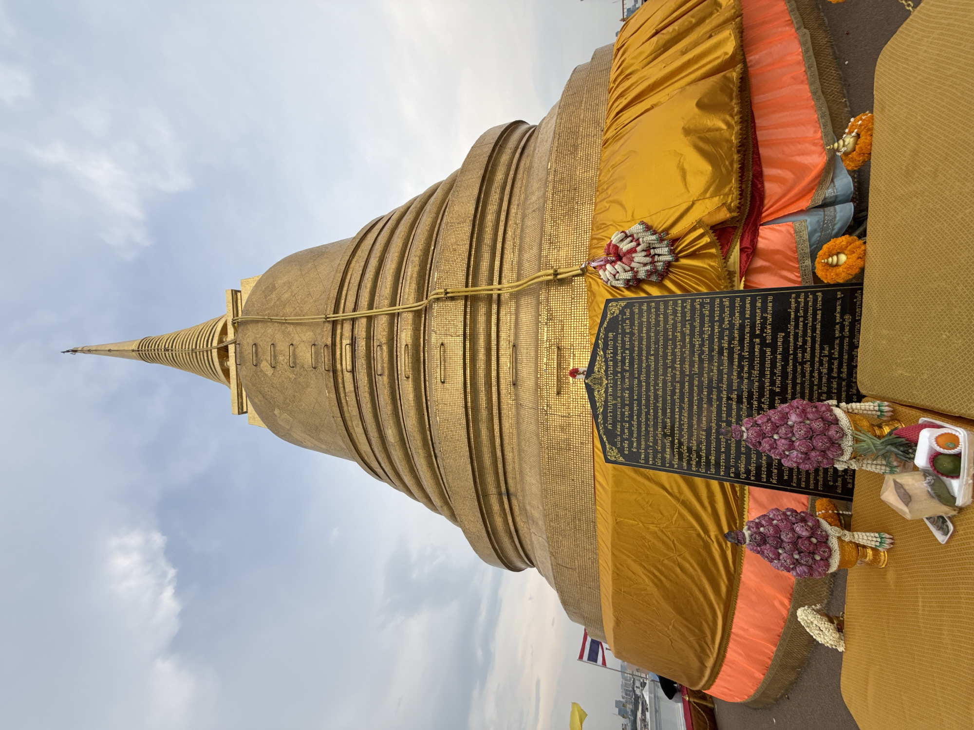 The image depicts Wat Saket, a prominent Buddhist temple complex in Bangkok, Thailand. The scene focuses on the Golden Mount (Phu Khao Thong), a gilded chedi. The chedi is covered in gold tiles and adorned with gold cloth and floral garlands. A black sign with Thai script stands to the side. The sky above is cloudy and pale blue. The edge of a city skyline can be seen in the far background.