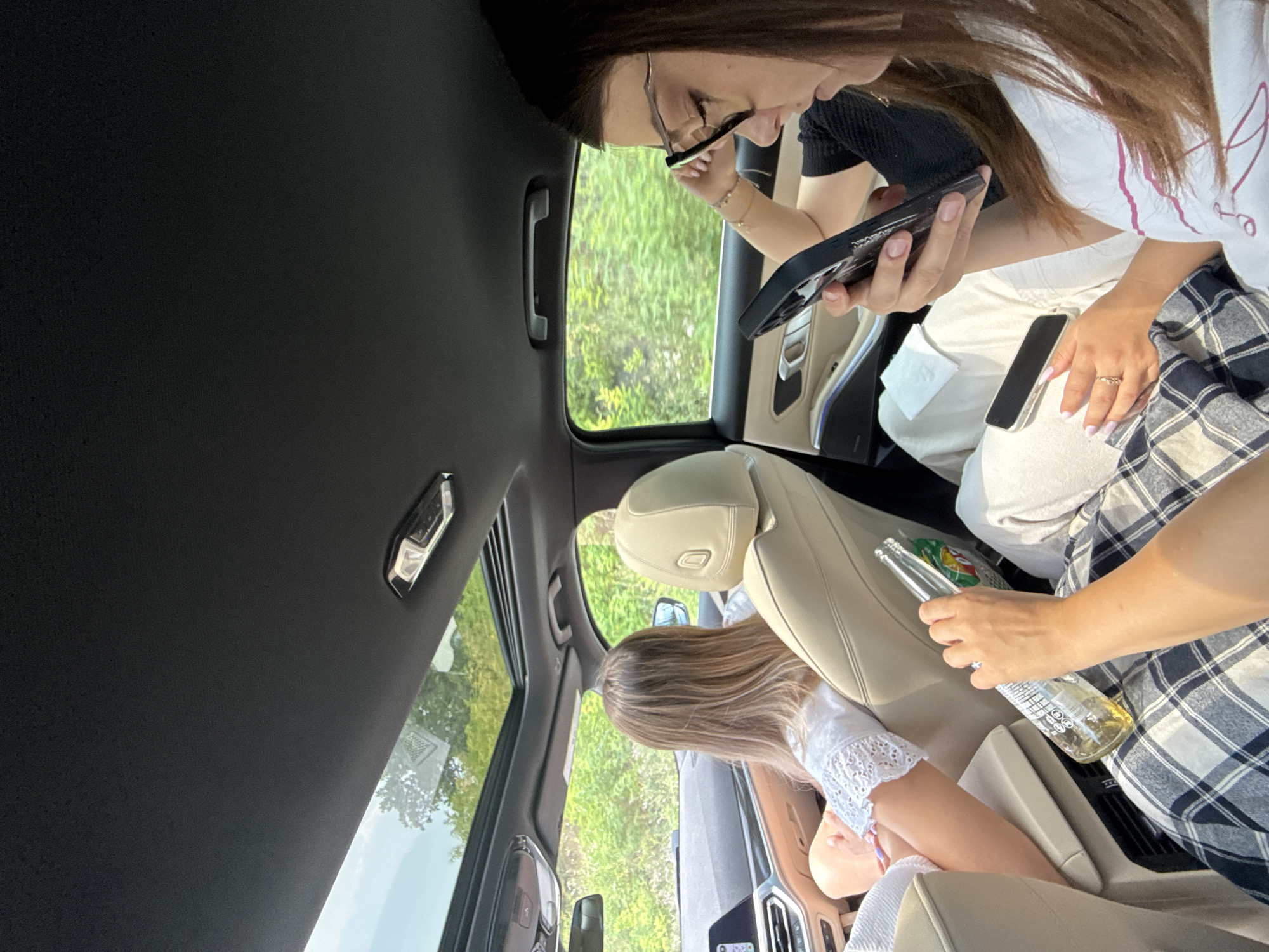The image depicts the interior of a car traveling on a road in the city of Rusca, Moldova. In the foreground, a person's arms are visible, holding a glass bottle of beer and resting on a checkered plaid fabric. To the left, a woman with blonde hair is seated in the driver's seat, looking forward. In the middle ground, a woman wearing glasses is looking down at her phone, her hand resting on her head. The car has a sunroof, and through the windows, lush green trees and vegetation are visible, indicating an outdoor, possibly rural or suburban, setting. The overall mood suggests a casual journey.