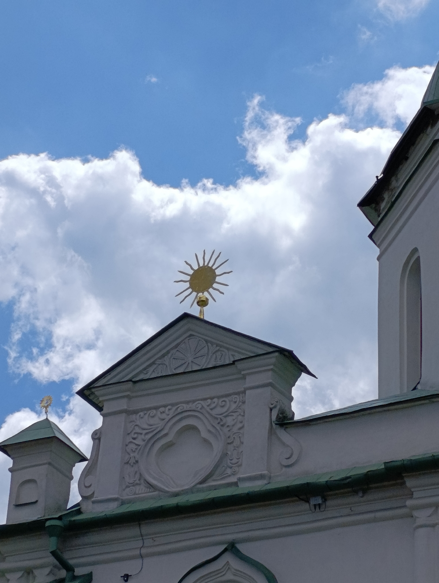 The image is a low-angle, outdoor photograph of the exterior of a white building, likely a church or historical structure, against a bright blue sky filled with fluffy white clouds. The building features ornate architectural details, including decorative moldings, carved patterns, and a pediment topped with a golden sunburst emblem. A smaller, similar emblem is visible on a smaller tower to the left. The roofline is visible with green tiling and some elements of water drainage. The lighting suggests it is daytime, with clear weather and bright sunshine casting subtle shadows. The image appears to be taken in Kyiv, Ukraine, given the architectural style often associated with Ukrainian religious buildings and the information provided. No people are visible in the frame. The overall mood is serene and majestic, highlighting the architectural beauty of the building against the backdrop of a pleasant day. There is no visible text in the image.