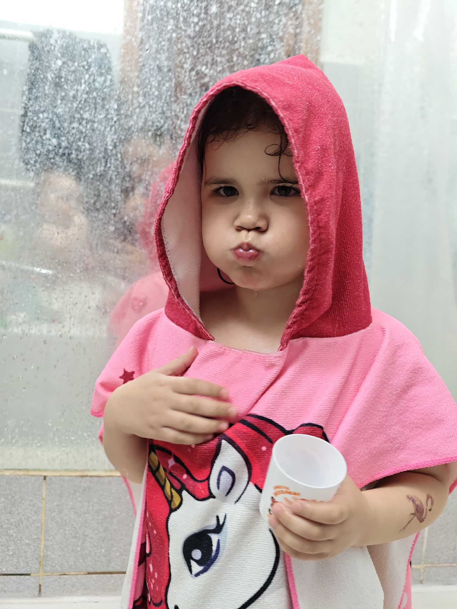 A young child, likely female, stands indoors, possibly in a bathroom, wearing a pink hooded towel with a unicorn design. The child is holding a white plastic cup in their right hand and has their left hand on their chest. The child has a temporary tattoo of a flamingo on their left arm. The child's face is expressive, with pursed lips. Behind the child is a shower door or enclosure covered in water droplets, reflecting a blurred image. The bathroom has light-colored tiles. The location is Chaikovsky, Russia.