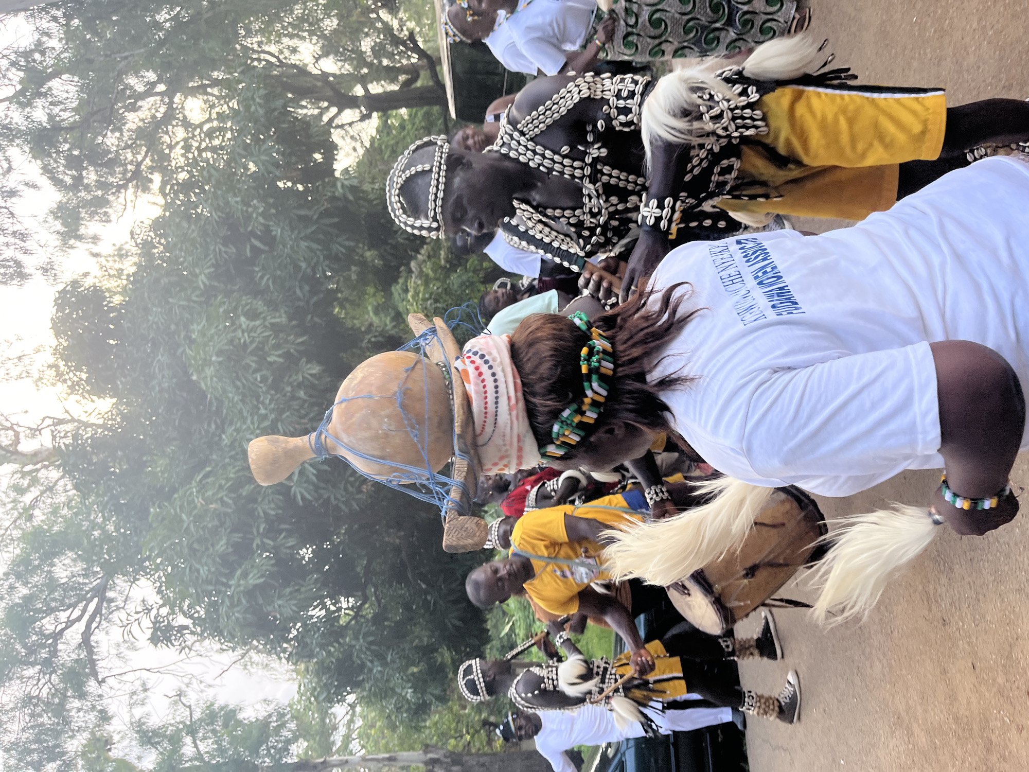 In an outdoor scene in Jos, Nigeria, a group of people are participating in a cultural event, likely a dance or ceremony. The foreground features two individuals in traditional attire. One person, facing away from the camera, wears a white t-shirt with blue text and a beaded headdress, holding a white feathered fan. To their right, a person in elaborate black and white beaded costume, including a headdress and chest adornment, is playing a drum. This individual wears yellow shorts and has white fur-like adornments on their arms and legs.
Behind them, other participants are visible. To the left, a person in a yellow t-shirt is also engaged with a drum. Further left, another individual in traditional attire with a beaded headdress and white and black striped attire holds a stick-like instrument. Several other individuals in the background are observing or participating in the event, some wearing white shirts and others in colorful patterned clothing. The environment is characterized by lush green trees and dappled sunlight filtering through the foliage, suggesting a daytime setting. A green car and a portion of a van are visible on the left and right sides of the frame, indicating the event is taking place in a somewhat urban or accessible outdoor area. The ground appears to be a dirt or gravel surface.