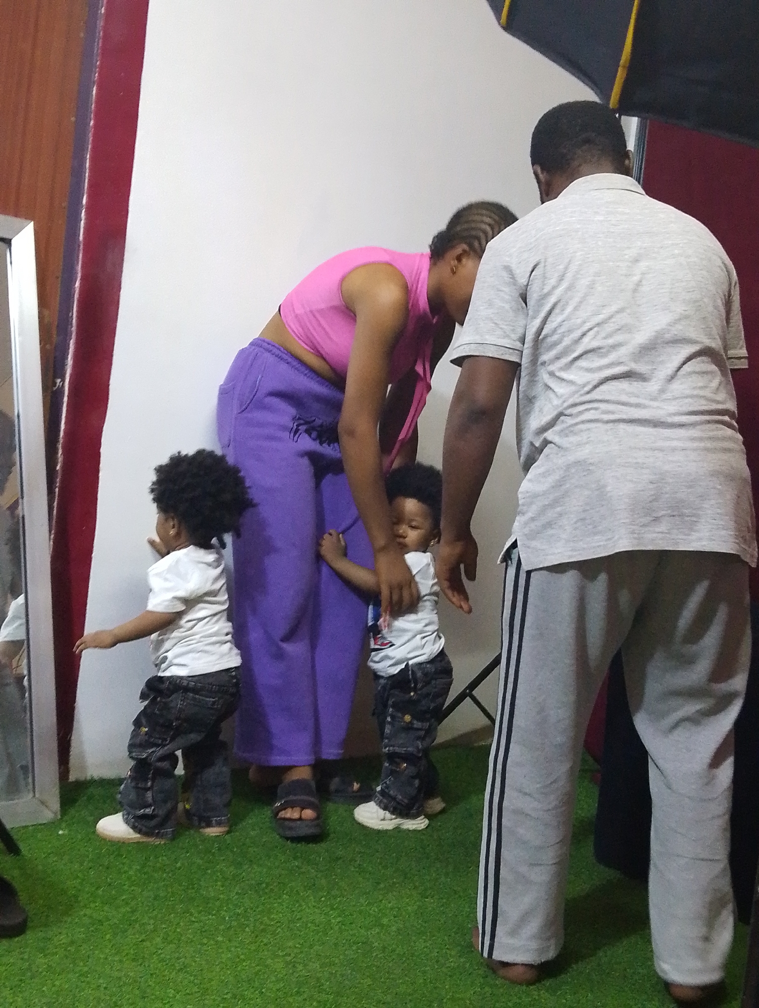 In an indoor studio in Babban Giji, Nigeria, two infants, a woman, and a man are present. The infant on the left wears a white shirt and dark denim-like pants with light-colored shoes, and has a large Afro hairstyle. The infant on the right wears similar clothing and footwear and is being held by the woman. The woman is wearing a pink crop top and purple sweatpants. The man stands on the right, wearing a grey t-shirt and grey sweatpants with black stripes. The background consists of a white wall with a red border on the left, a mirror, artificial green turf flooring, and an open black photography umbrella.