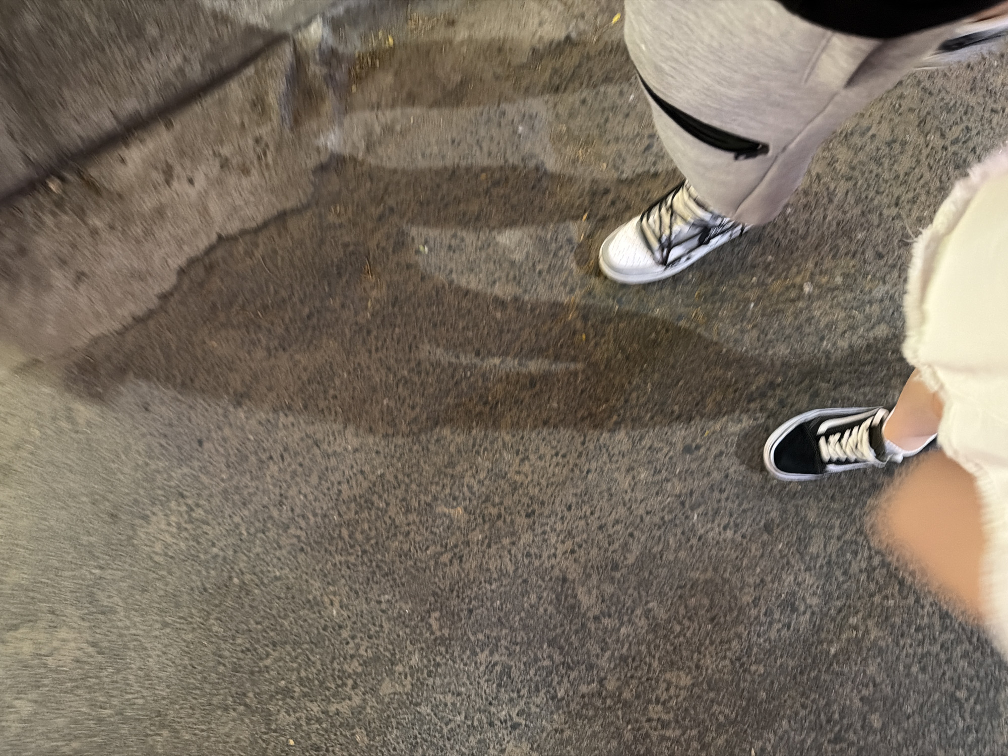 The image depicts a street scene, possibly in Đà Nẵng, Vietnam, based on the appearance of the asphalt and sidewalk. Two individuals are partially visible: one wearing grey sweatpants and white sneakers, the other wearing white shorts and black Vans sneakers. The ground is damp, reflecting light in patches, suggesting recent rain or cleaning. The focus is on the street surface and the lower portions of the individuals. No specific actions or interactions are discernible.