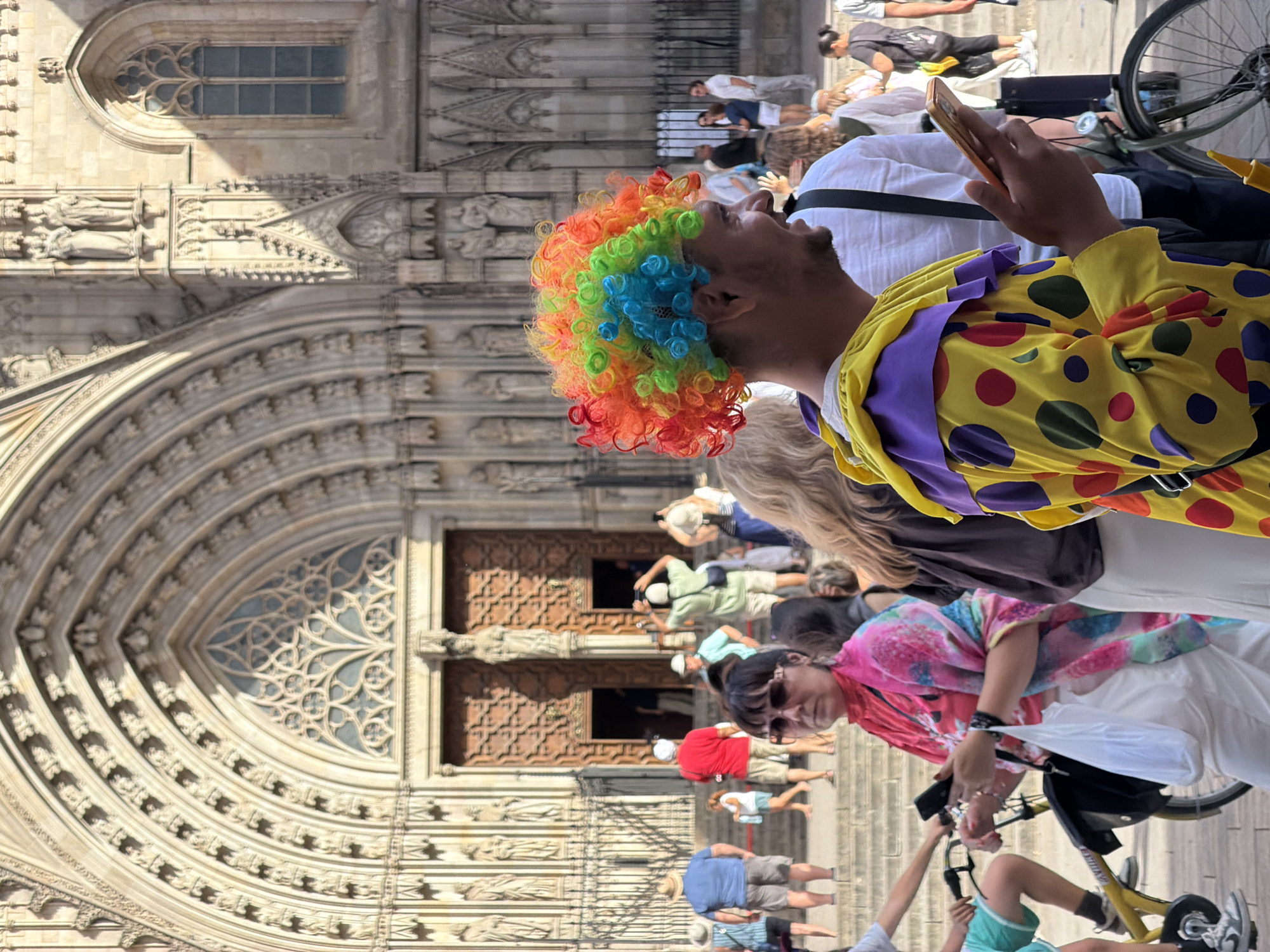 In front of the Barcelona Cathedral, a man in a colorful clown wig and costume is holding a smartphone. He is smiling and looking to his right, with a crowd of people behind him. The cathedral's facade is visible in the background, with its intricate stone carvings and large wooden doors. Sunlight casts shadows on the building, indicating it is daytime. Other people in the image are dressed casually, and some are holding cameras or phones, suggesting they are tourists. A bicycle is parked to the right of the clown. The scene appears to be a lively public gathering.