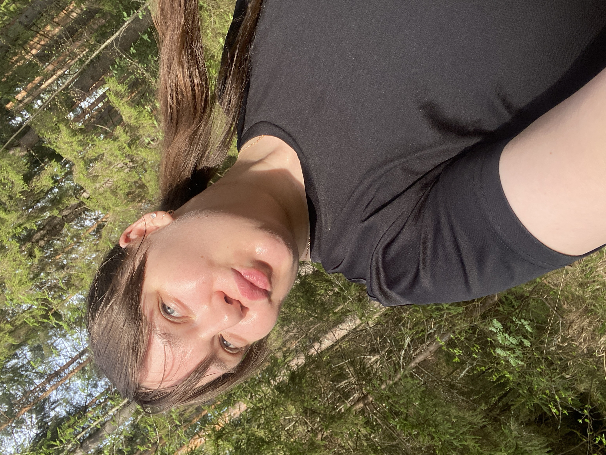 A young, fair-skinned person with brown hair is the central subject of the image. They wear a black t-shirt. The background suggests an outdoor setting, possibly a wooded area, with trees and greenery visible. Given the prompt's mention of Votkinsk, Russia, the location could be a park or forest area within or near the city. No specific buildings or landmarks identifiable to pinpoint an exact location are present in the image. The person is looking toward the camera and the image is turned 90 degrees to the left.